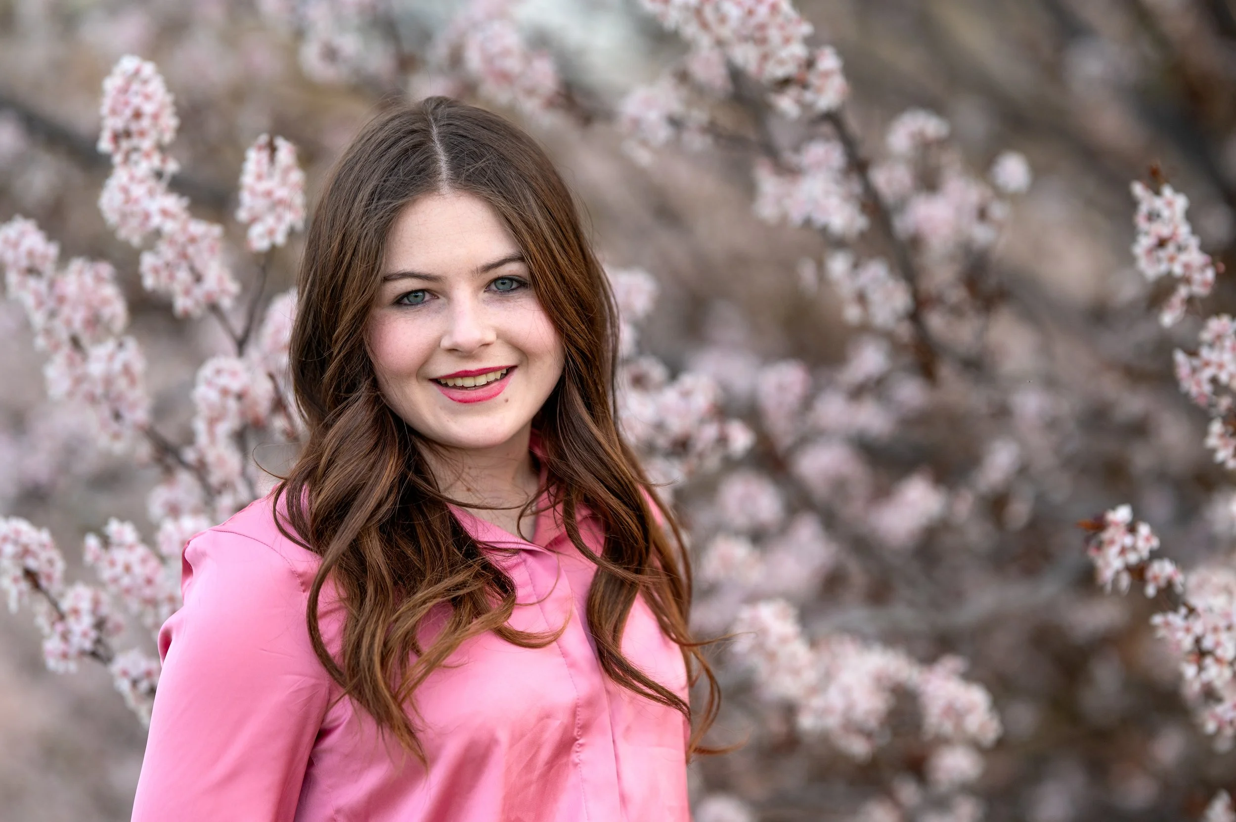 A smiling young brunette girl with a pink blouse in front of soft pink cherry blossoms in Murray Utah