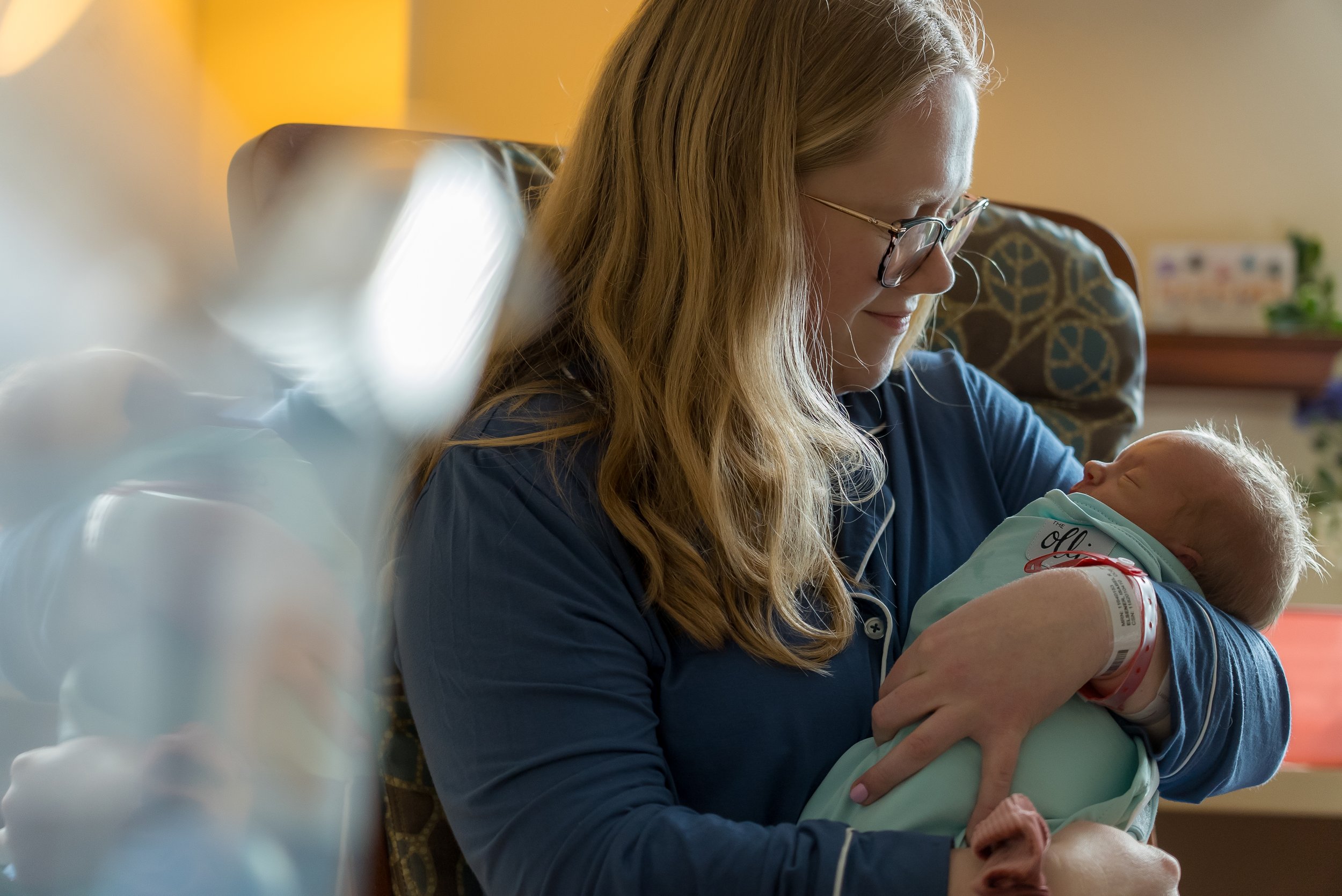 Golden Hour Photography Services: A woman holding a newborn baby while sitting on a chair, smiling and looking at the baby, in a warmly lit hospital room.