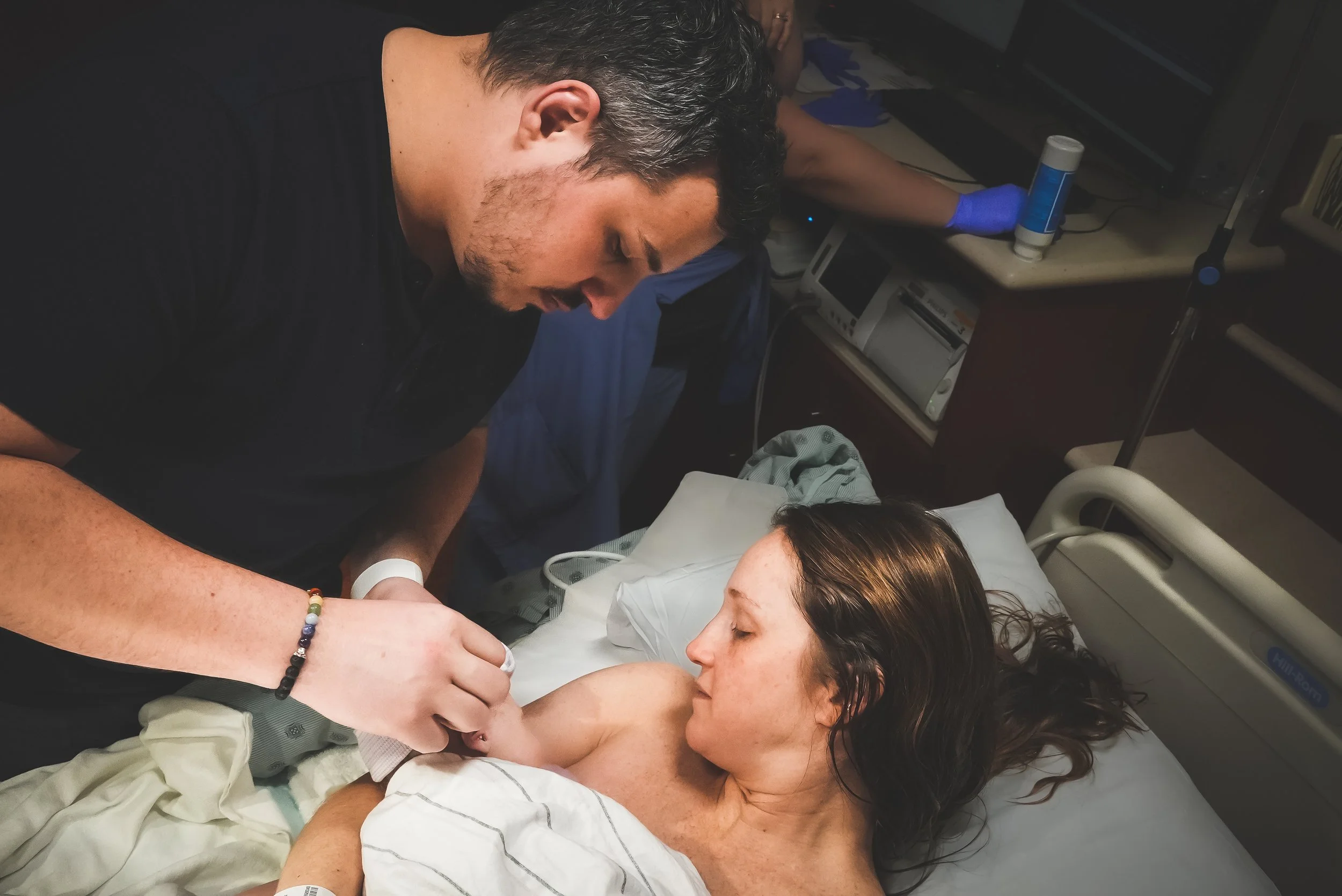 Birth Photography: A father in a black shirt leaning over a mother lying in a hospital bed, touching their newborn baby.