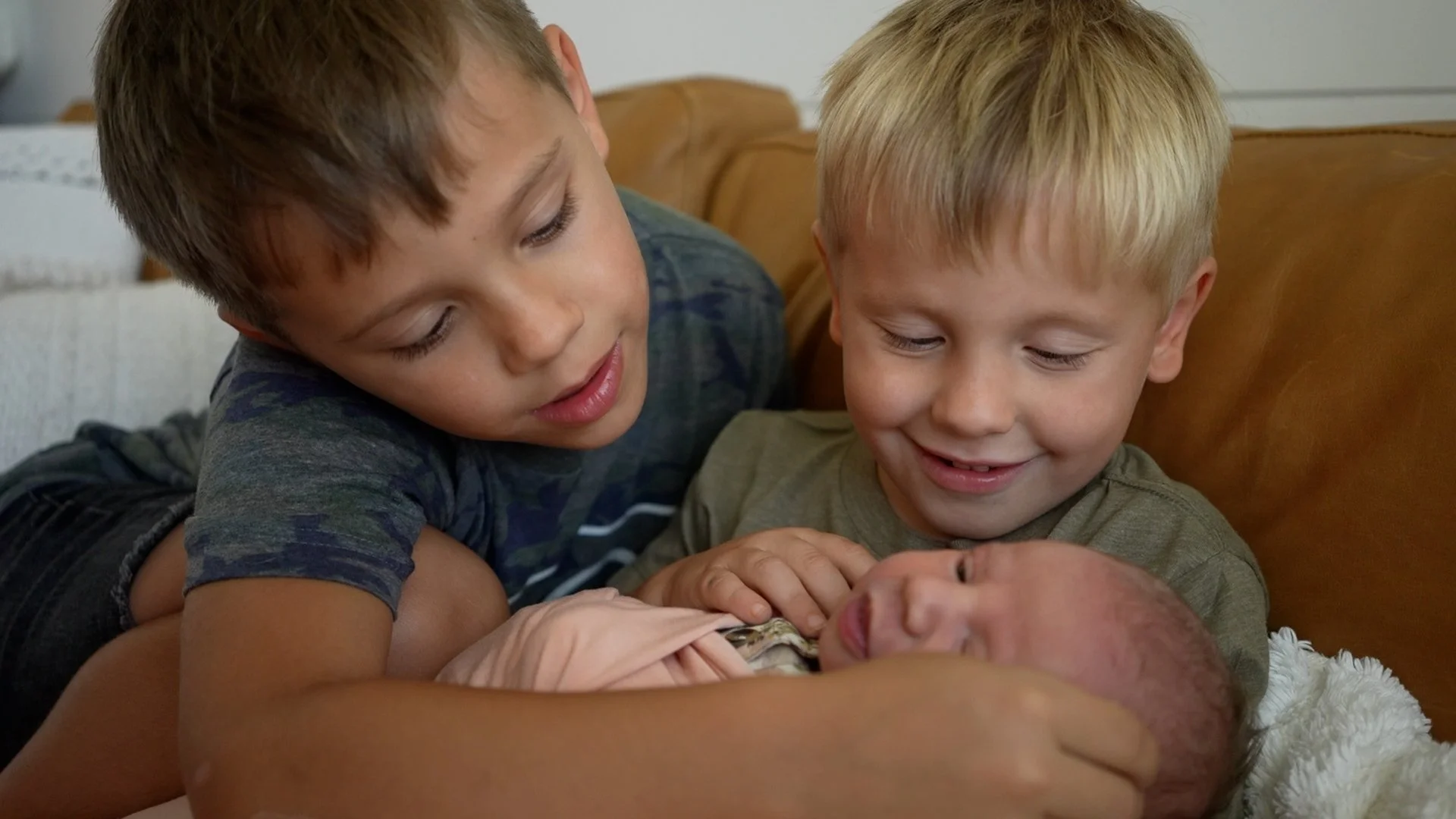 Homecoming Story Videography ServicesL Two young boys smiling and gathered around their newborn baby sister lying on a blanket.