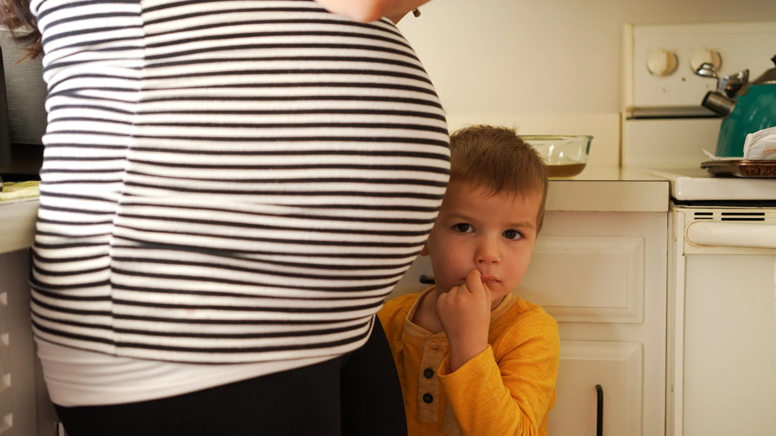 Family photography: A young boy with short brown hair, wearing a yellow shirt, standing in a kitchen with his mama's pregnancy belly, and looking at the camera while biting his thumb.
