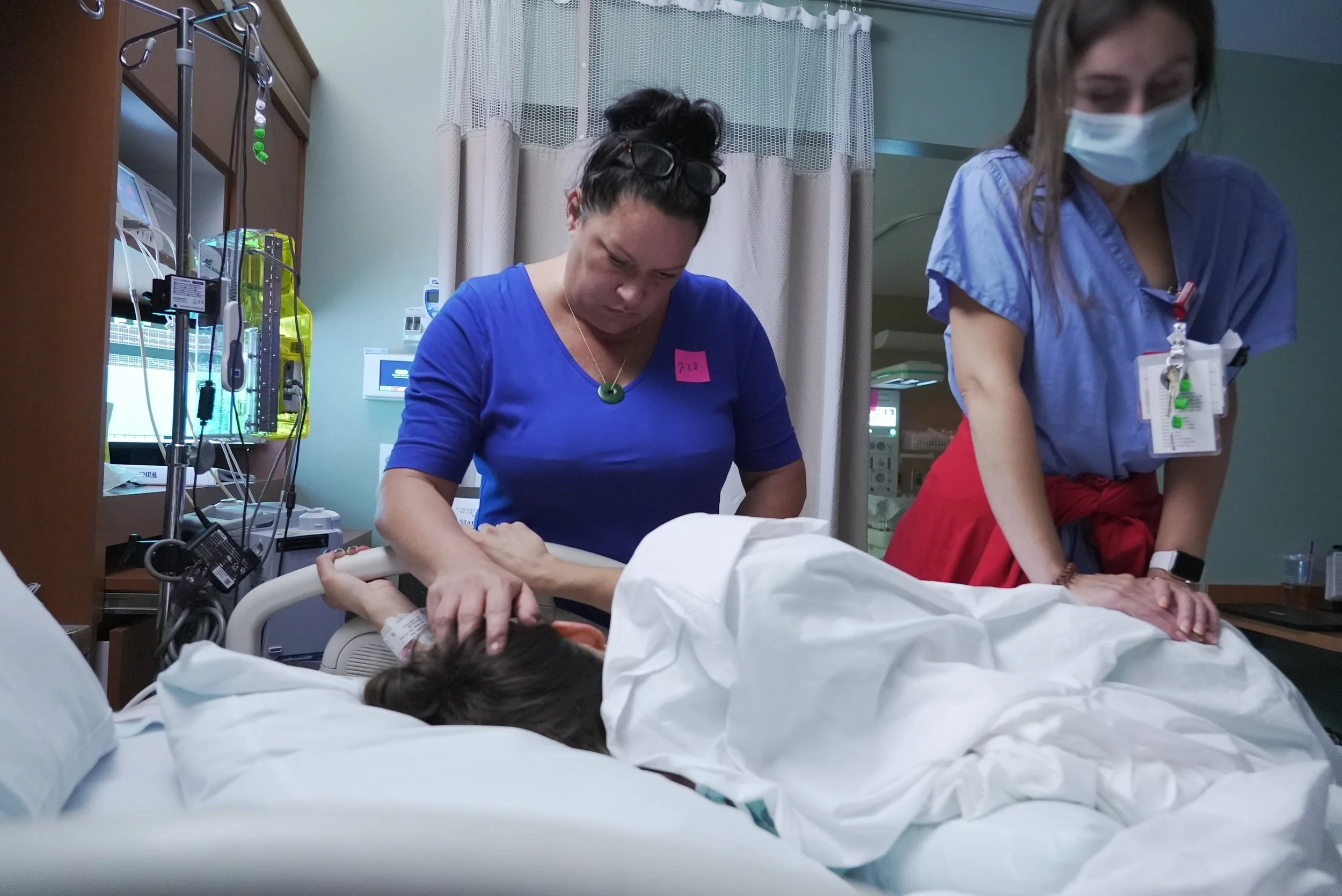 Birth Photography: A woman labors on her side in a hospital bed with her mom stroking her hair and a nurse providing counter-pressure.