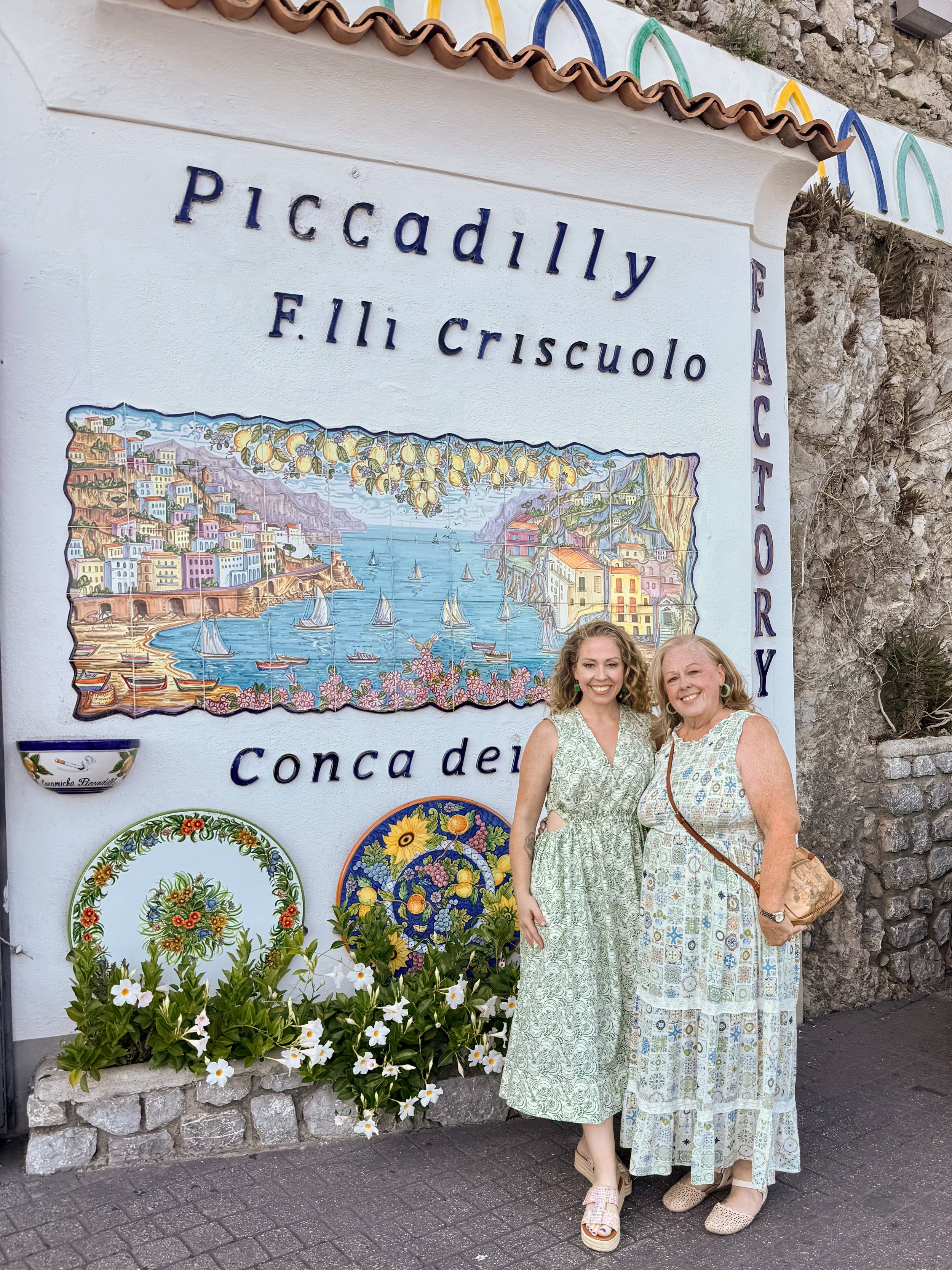 Certified Doula and photographer/videographer Nina Koehler and her mom standing in front of a painted sign for Piccadilly F.lli Criscuolo Conca dei Marini, with colorful flowers and decorative plates beneath it.