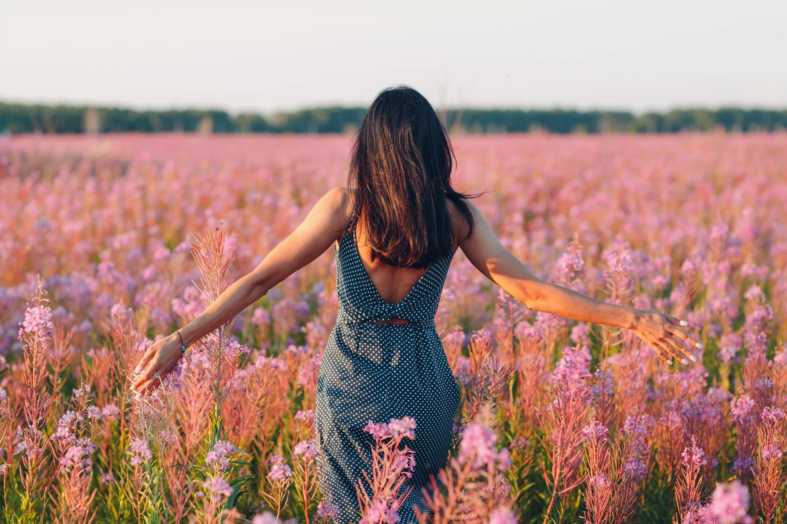 A woman with dark hair in a polka dot dress standing in a field of pink and purple flowers with arms outstretched.