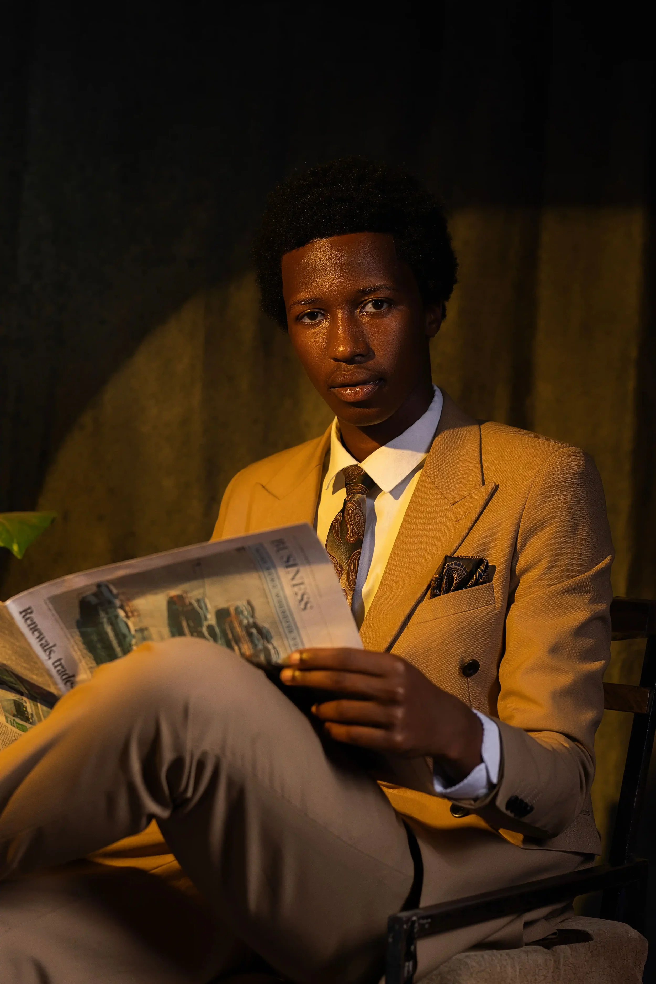 A person with dark skin and short curly hair, dressed in a tan suit, white shirt, and tie, is sitting and reading a newspaper in a dimly lit setting.