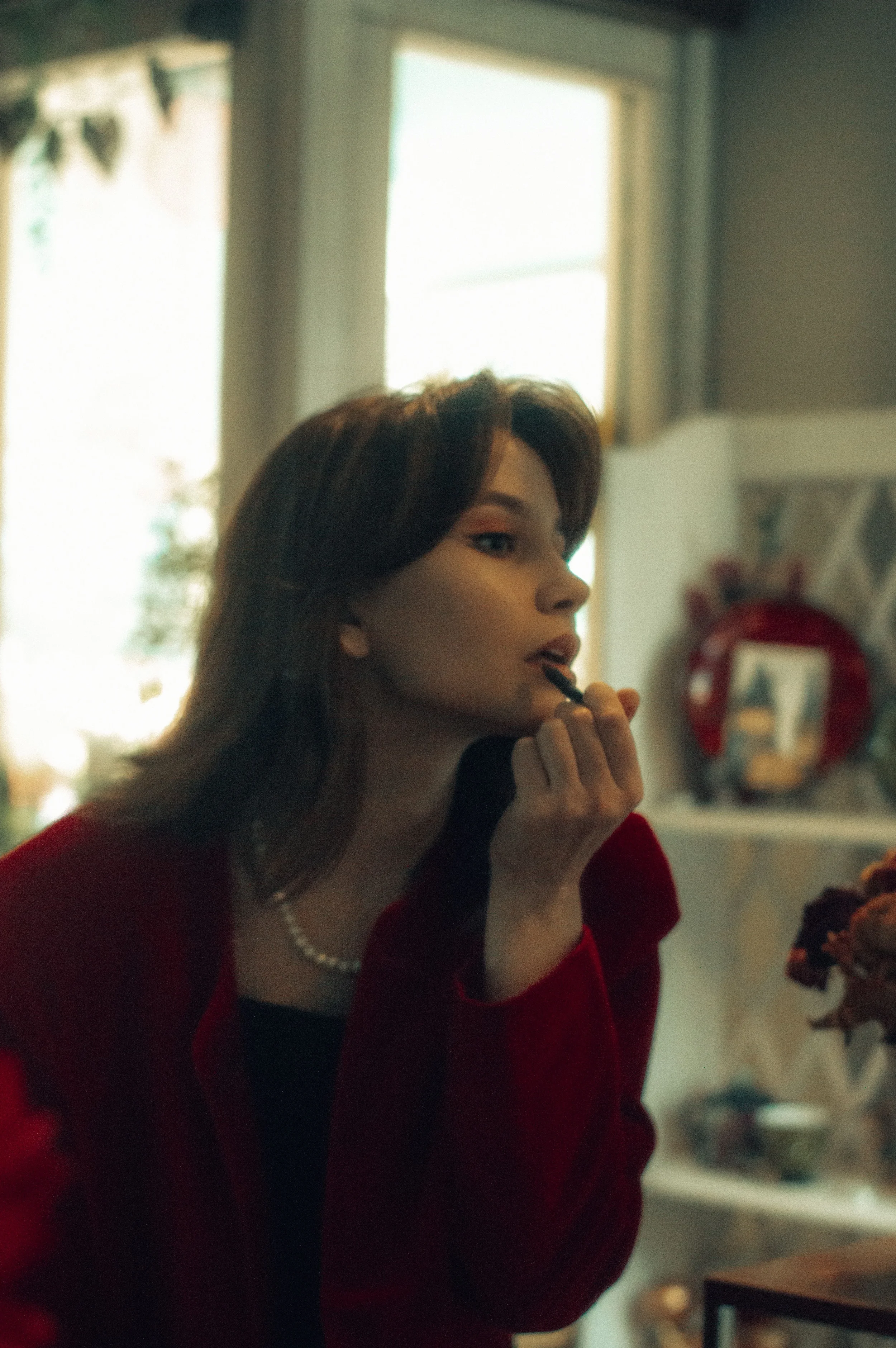 A woman with dark hair and a pearl necklace sitting indoors, applying lipstick, wearing a red and black outfit.