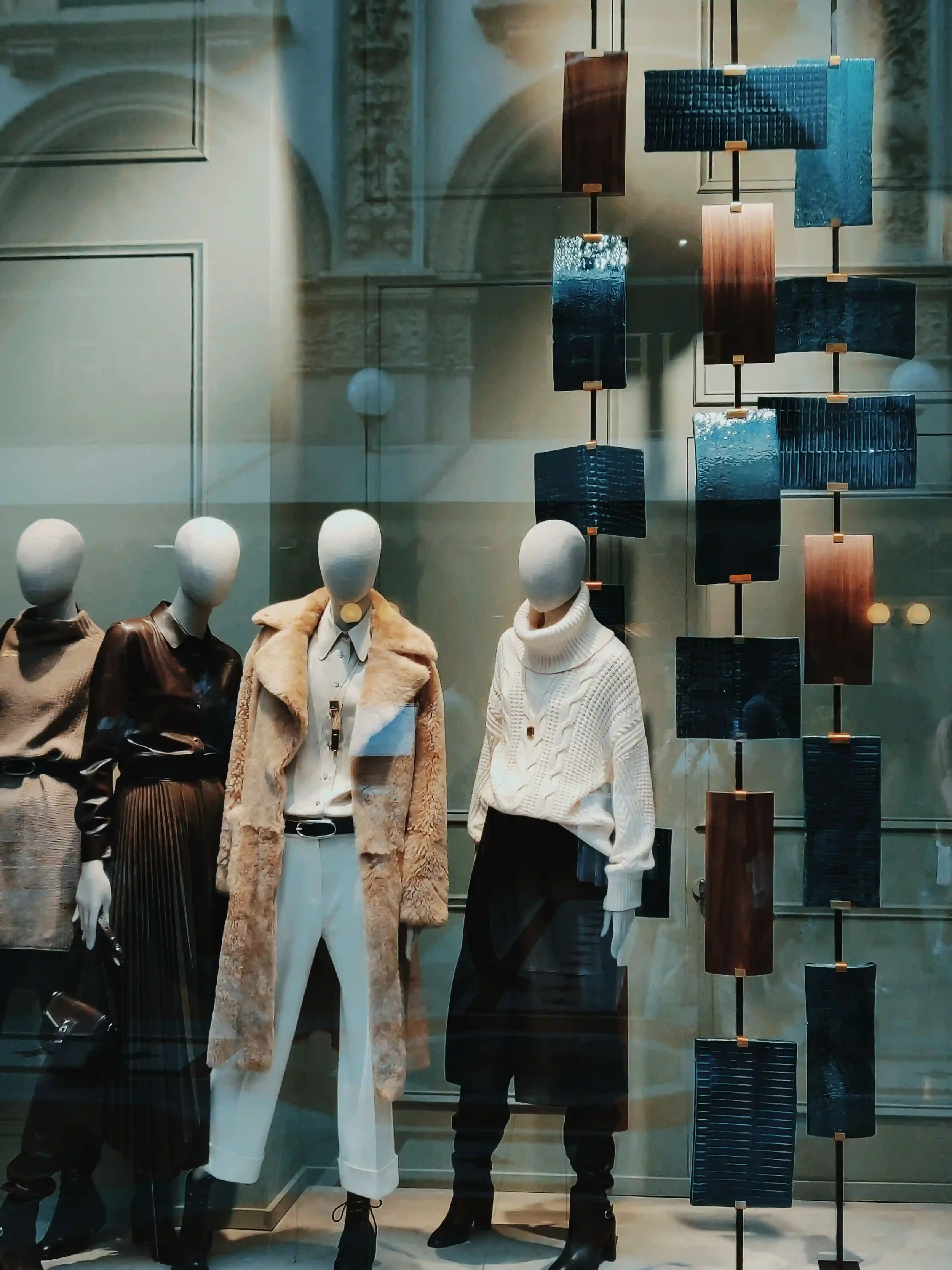 Display window with mannequins dressed in stylish winter clothing including sweaters, coats, and boots, along with a decorative art piece featuring rectangular panels in various colors and textures.