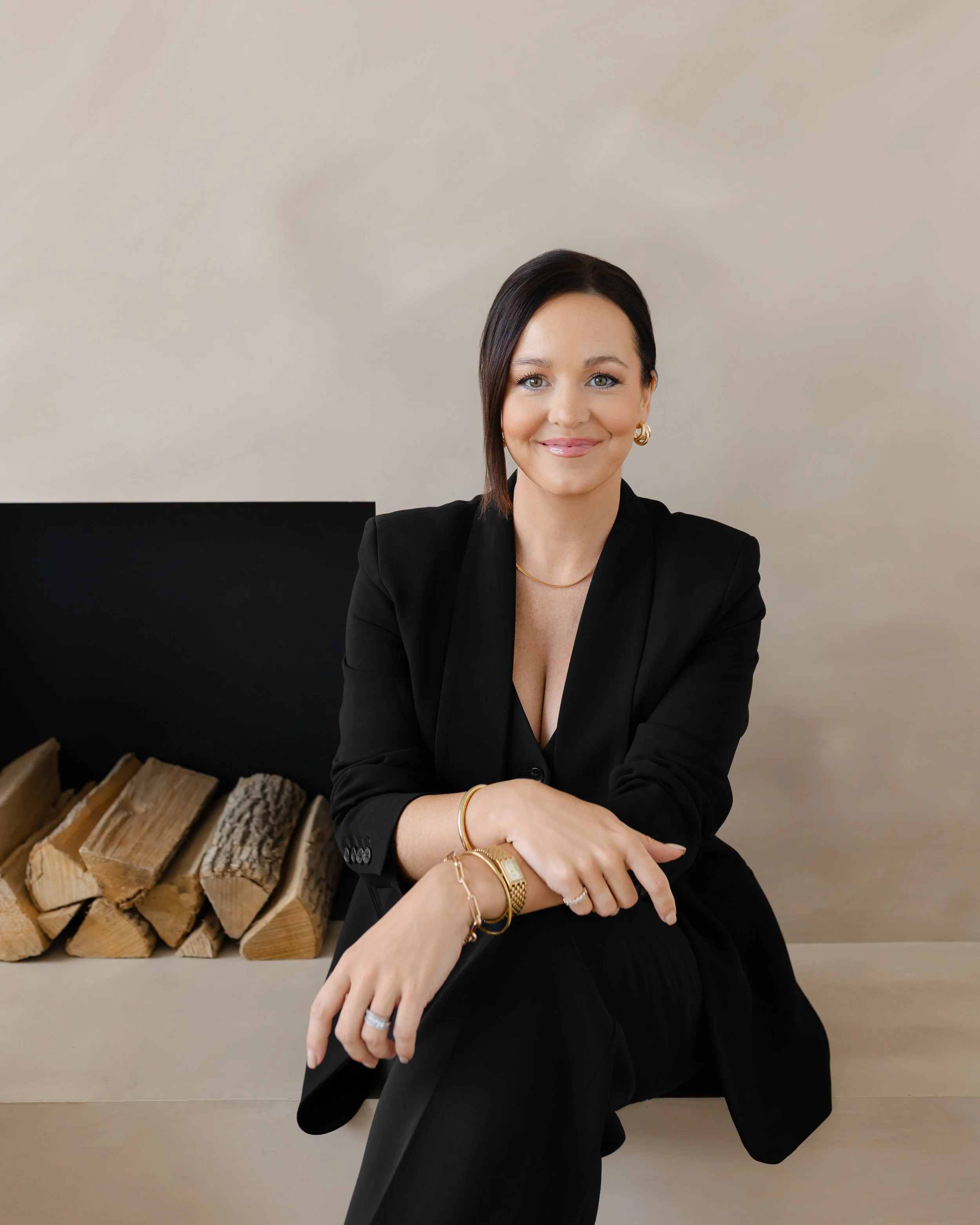 A woman with dark hair and light eyes, dressed in a black blazer, smiling while sitting with her hands resting on her crossed knee. In the background, there is a beige wall and a stack of firewood.