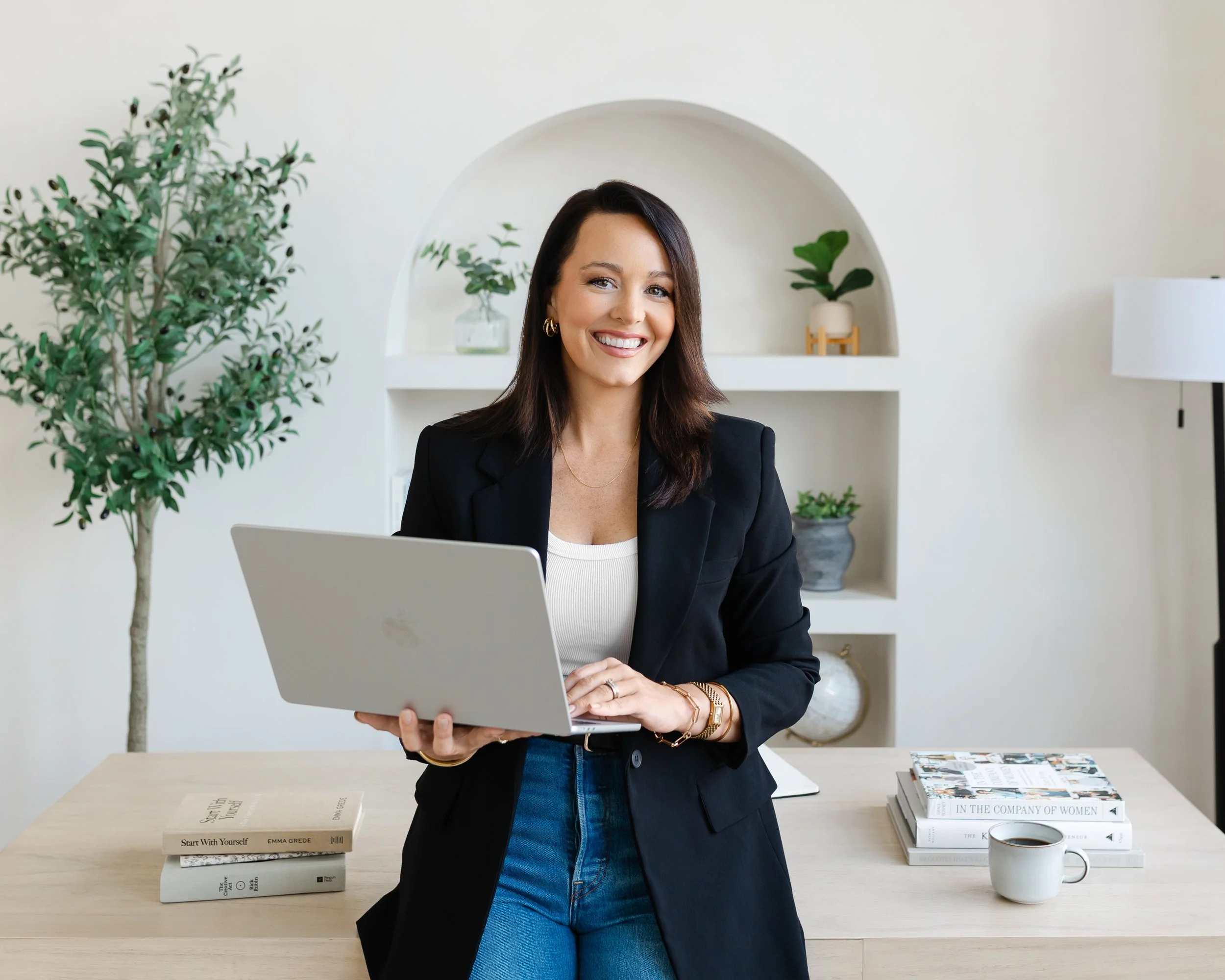 A smiling woman in a black blazer holding a laptop in a modern, minimalist office. Behind her are white shelves with plants, books, and magazines on a light wood desk.