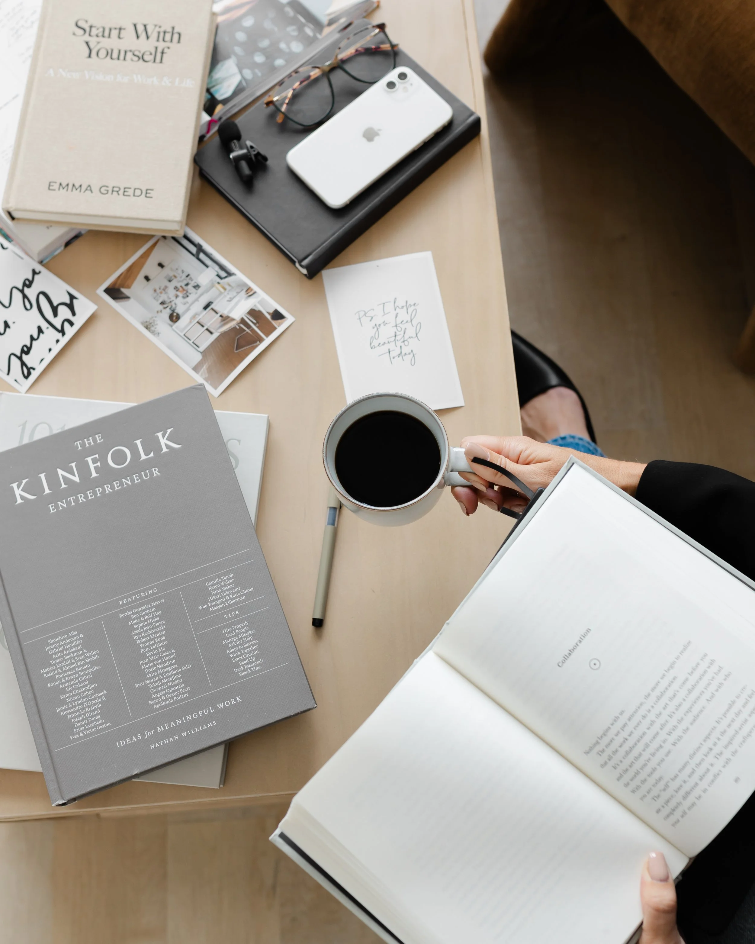 A cluttered desk with books, an iPhone, glasses, a notebook, a pen, a cup of coffee, and photographs. One book titled 'Start With Yourself' by Emma Grede is prominently visible.