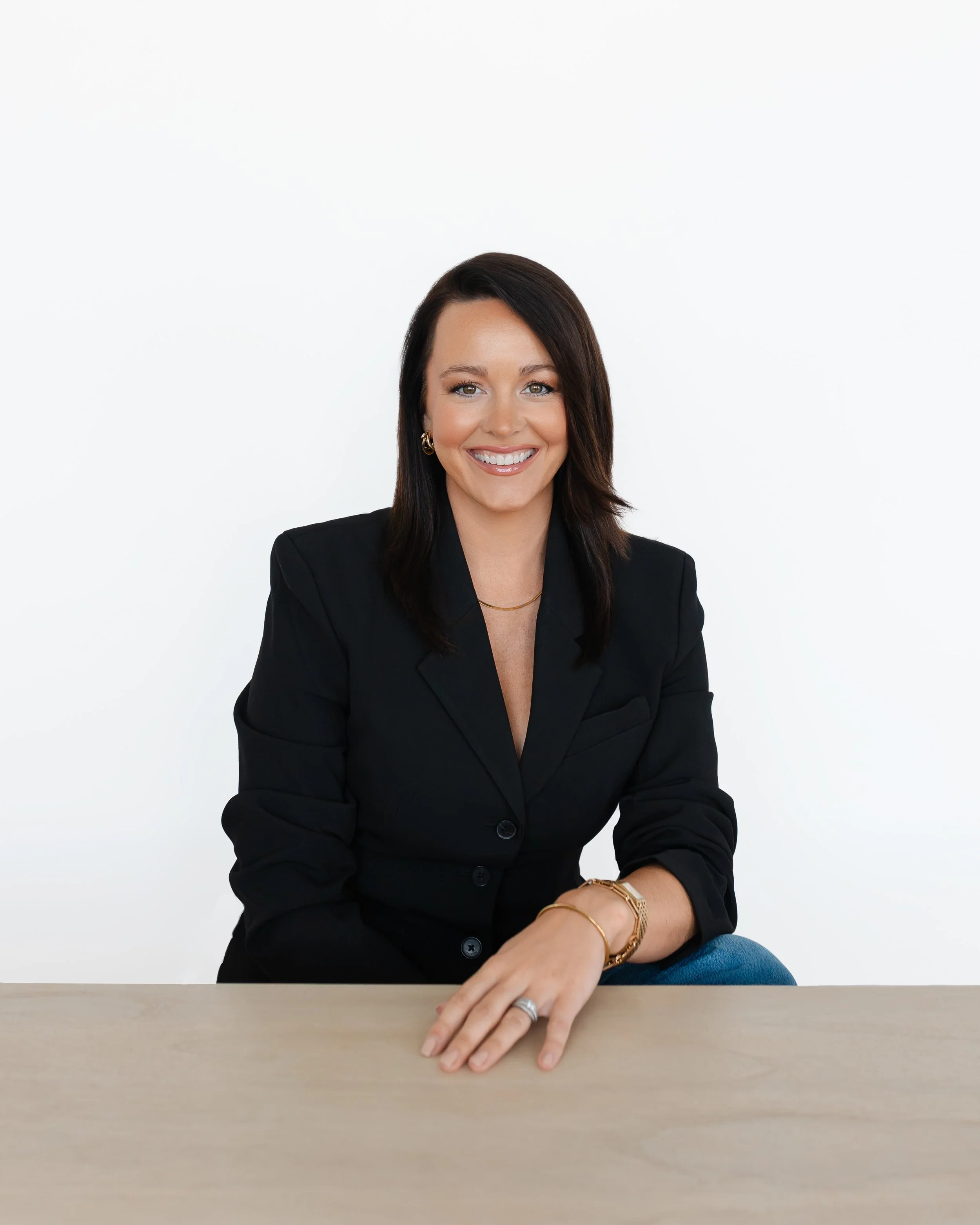 A woman with dark brown hair, wearing a black blazer, smiling, sitting at a light-colored table with her right arm resting on it and a gold bracelet on her wrist, in front of a plain white background.