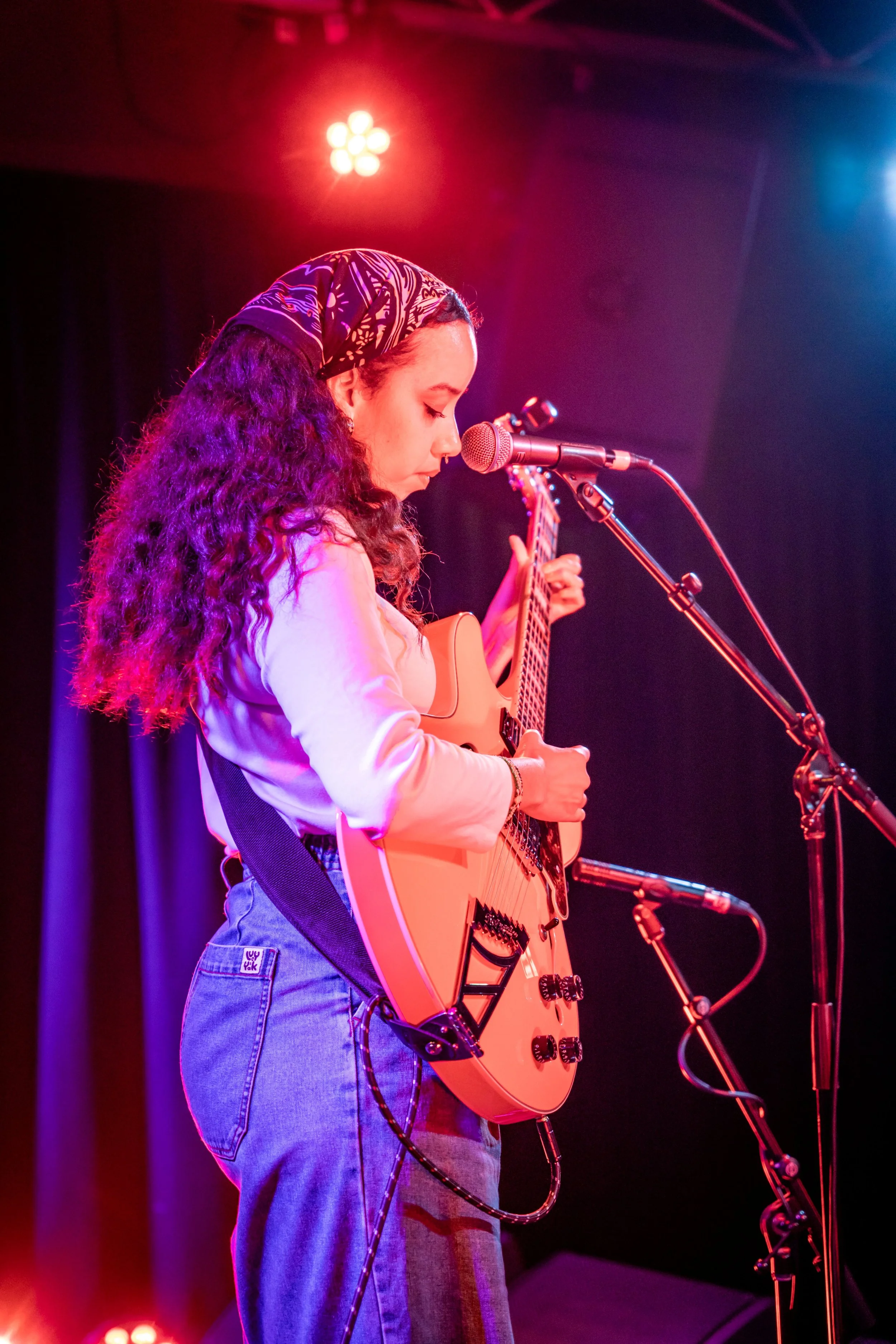 A woman with curly hair wearing a bandana and a cream-colored top, performing on stage with an acoustic guitar and singing into a microphone.