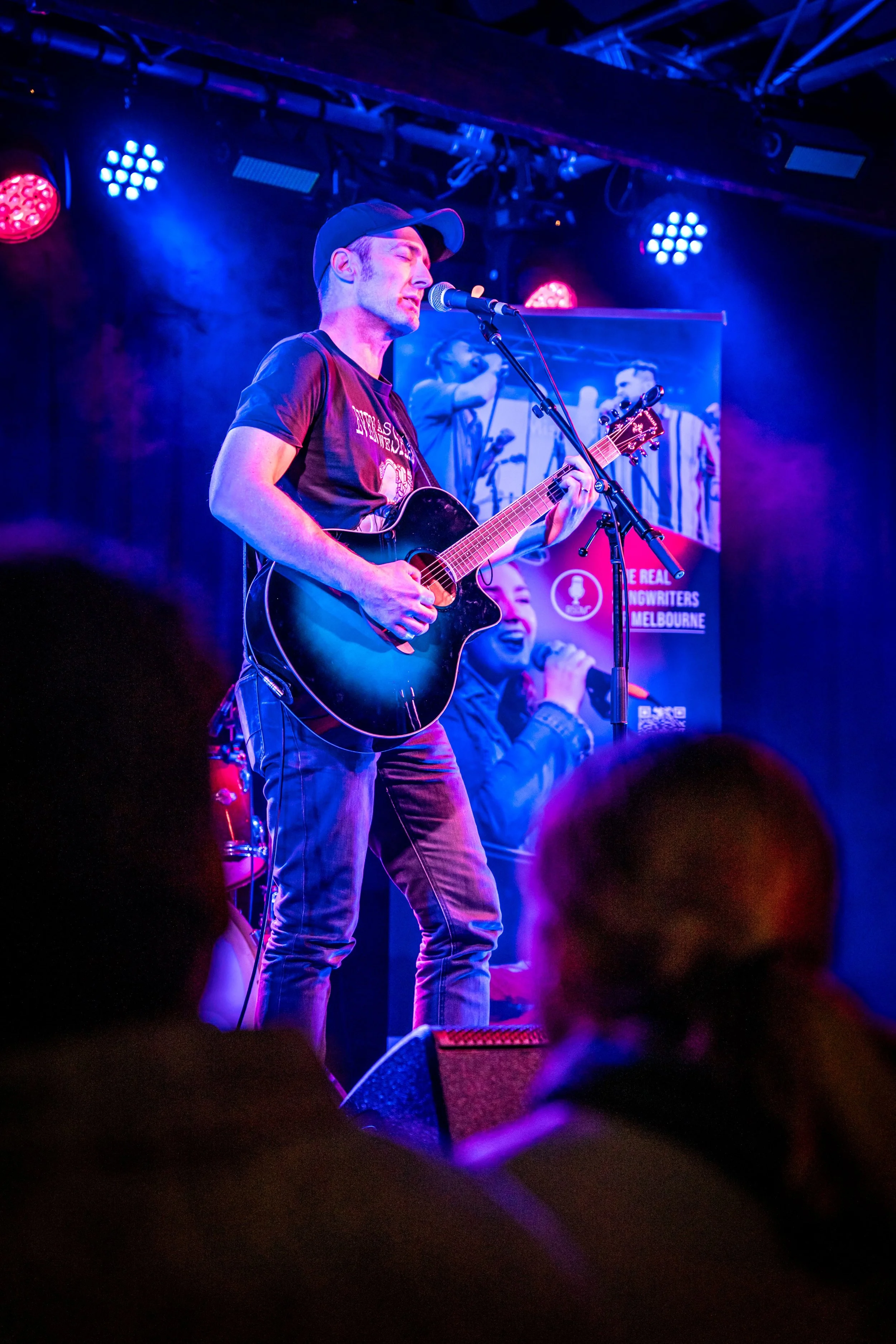 Musician performing with guitar on stage at a live concert, illuminated by colorful stage lights, with audience in foreground and a poster in the background.