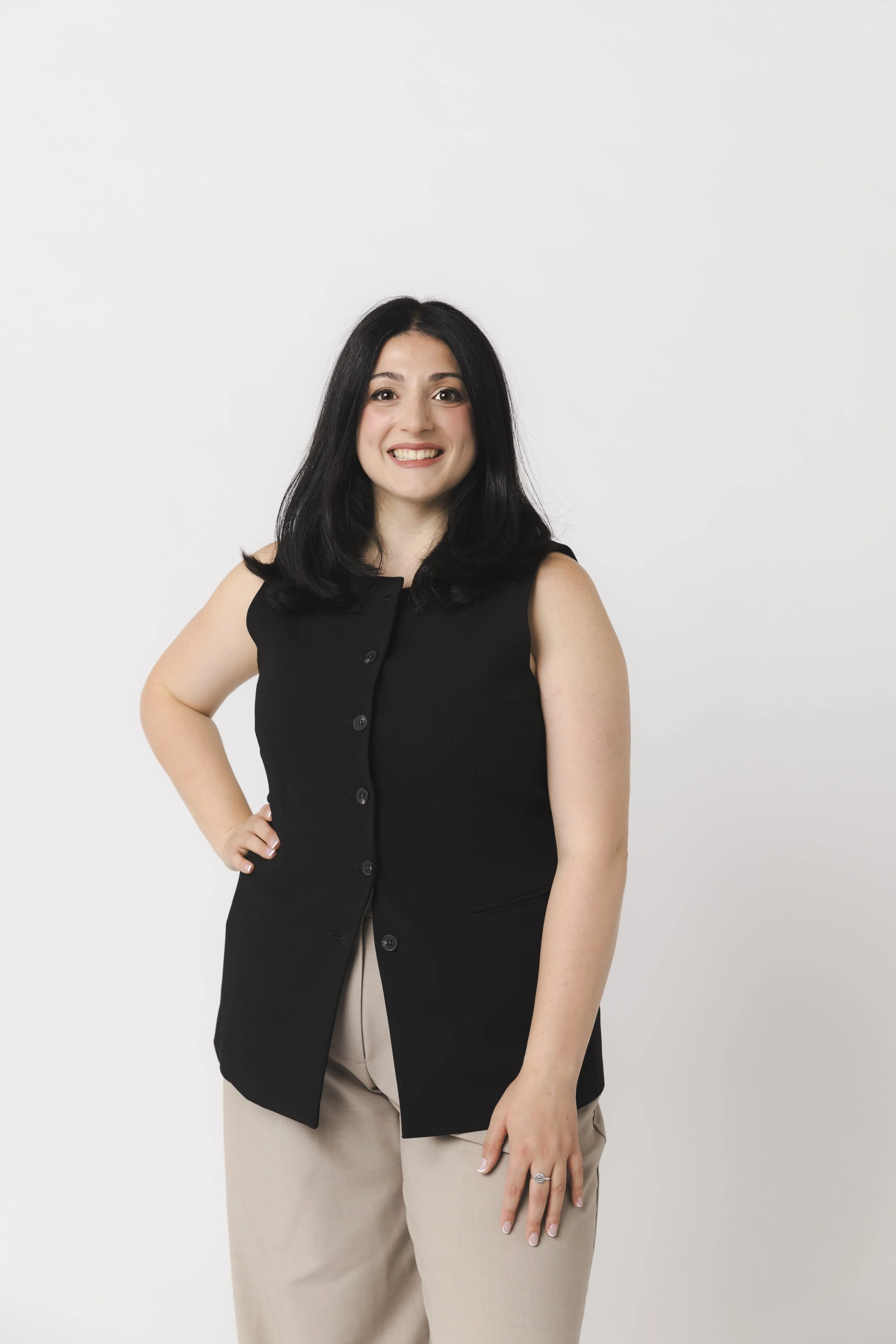 Smiling woman with black hair wearing a black sleeveless top and beige pants standing against a white background.
