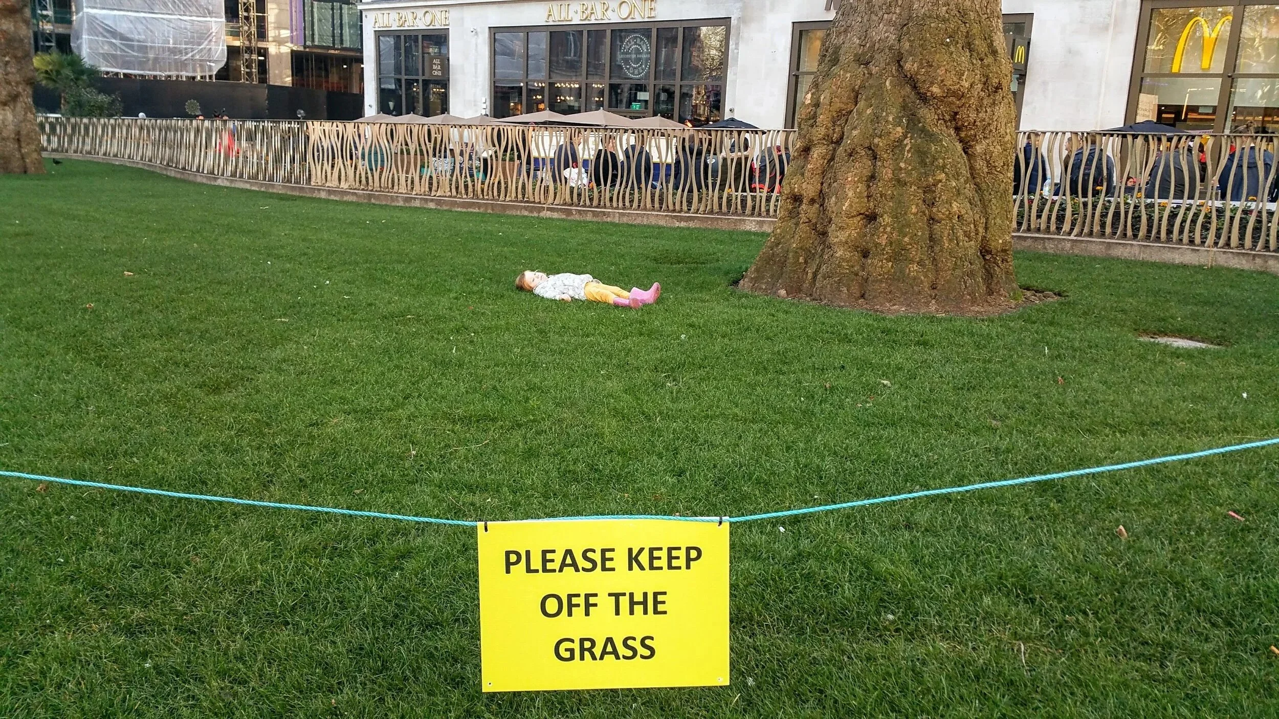 A young girl is lying on her back on the grass. The girl is not responding to her parents calling her off the grass. There is a yellow sign in the foreground that says, 'Please keep off the grass.'