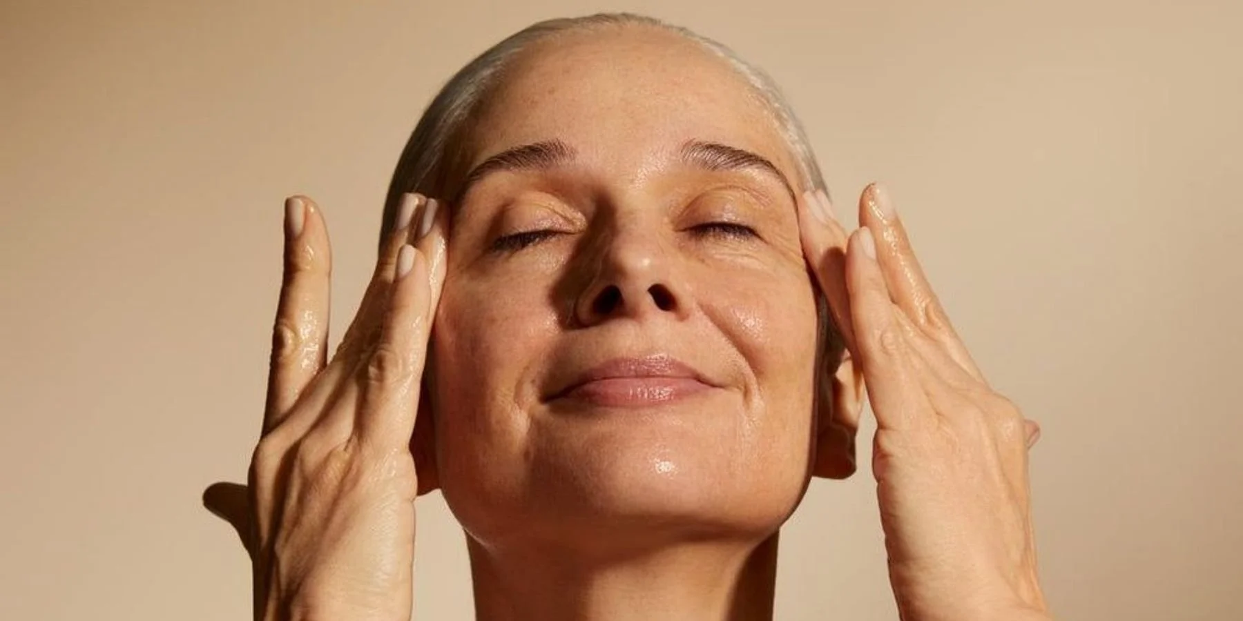 A menopausal woman touching her face with her eyes closed against a beige background