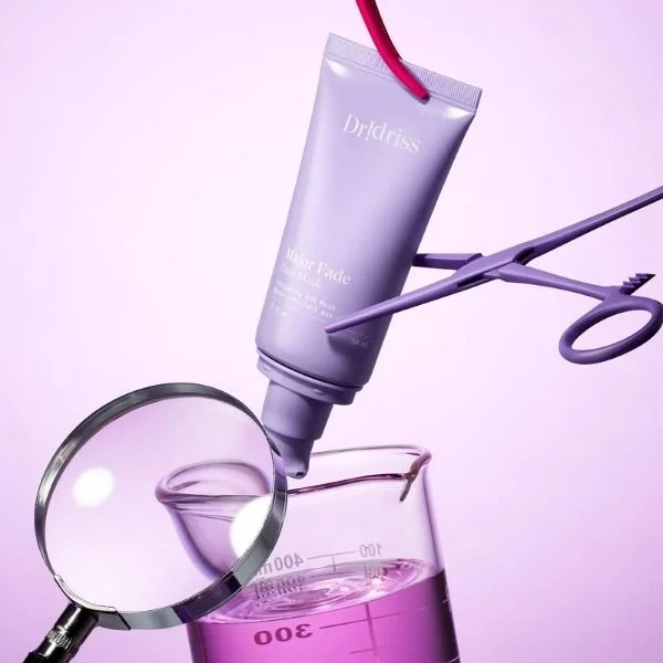 An image of a Dr Idriss face mask with a pair of scissors cutting it, a magnifying glass and a beaker of pink liquid against a lilac background