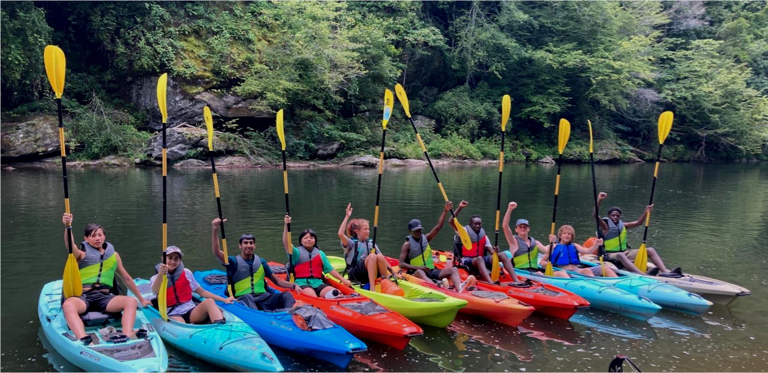 Group of pre-teens and teenagers on a river in kayaks raising their hands in the air.
