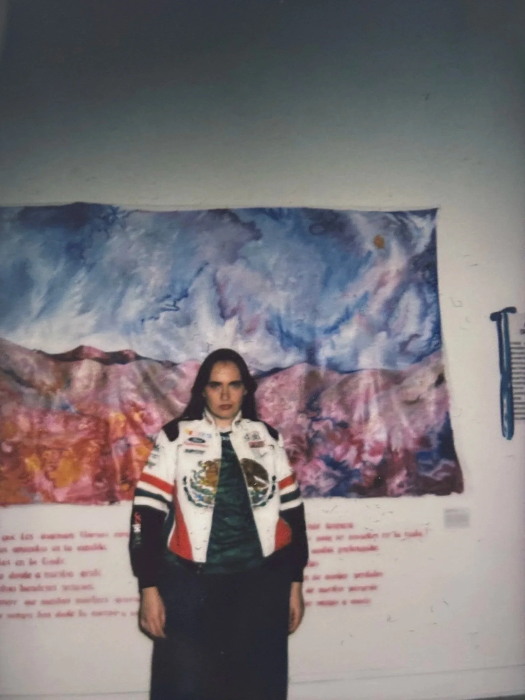 A woman in a black skirt and white jacket with colorful patches stands in front of a large backdrop with a cosmic landscape of mountains and sky.