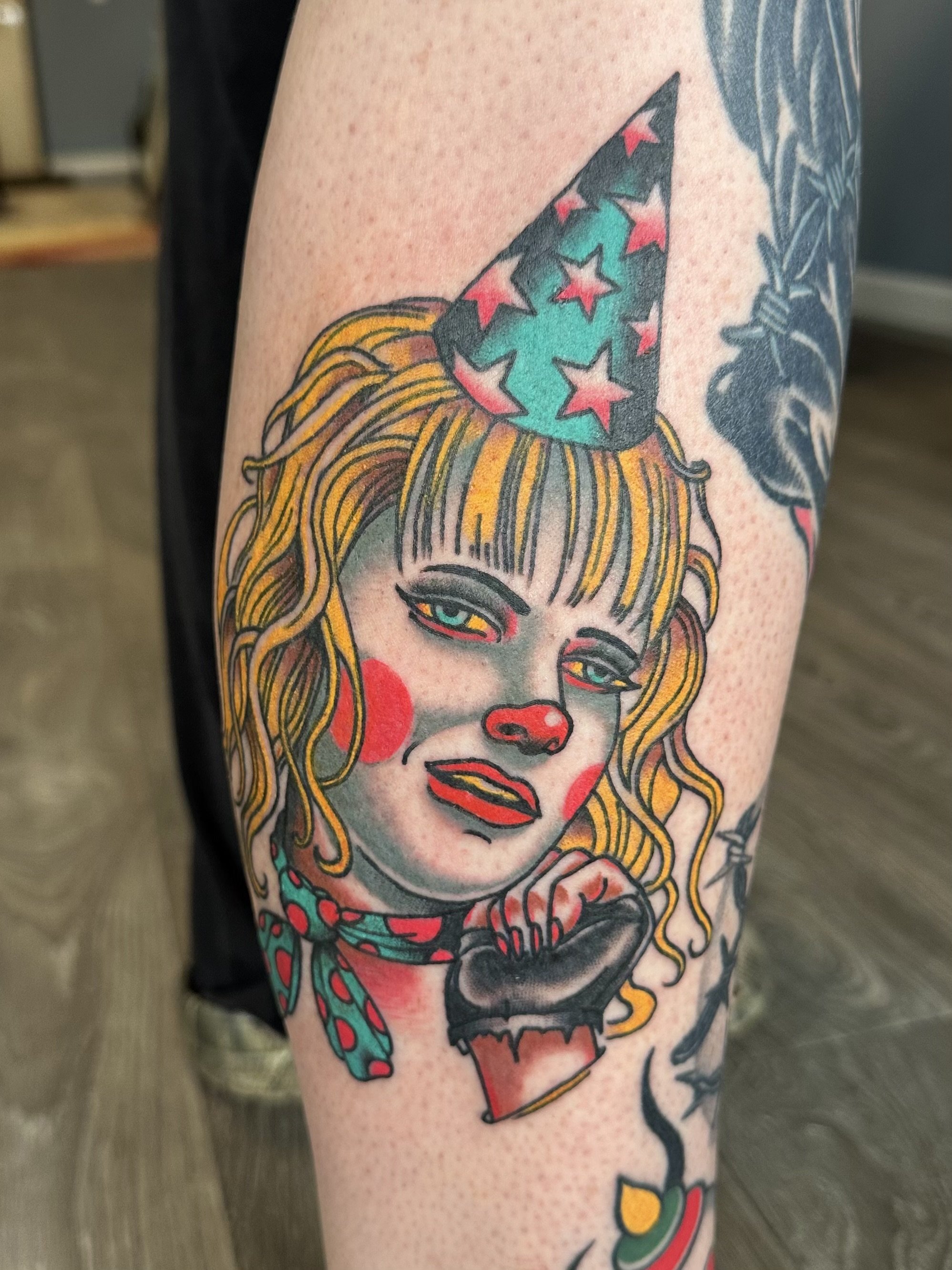 Colorful tattoo of a woman with blonde hair, red lipstick, and a blue eye, wearing a polka-dot dress, and a party hat with stars on her head.