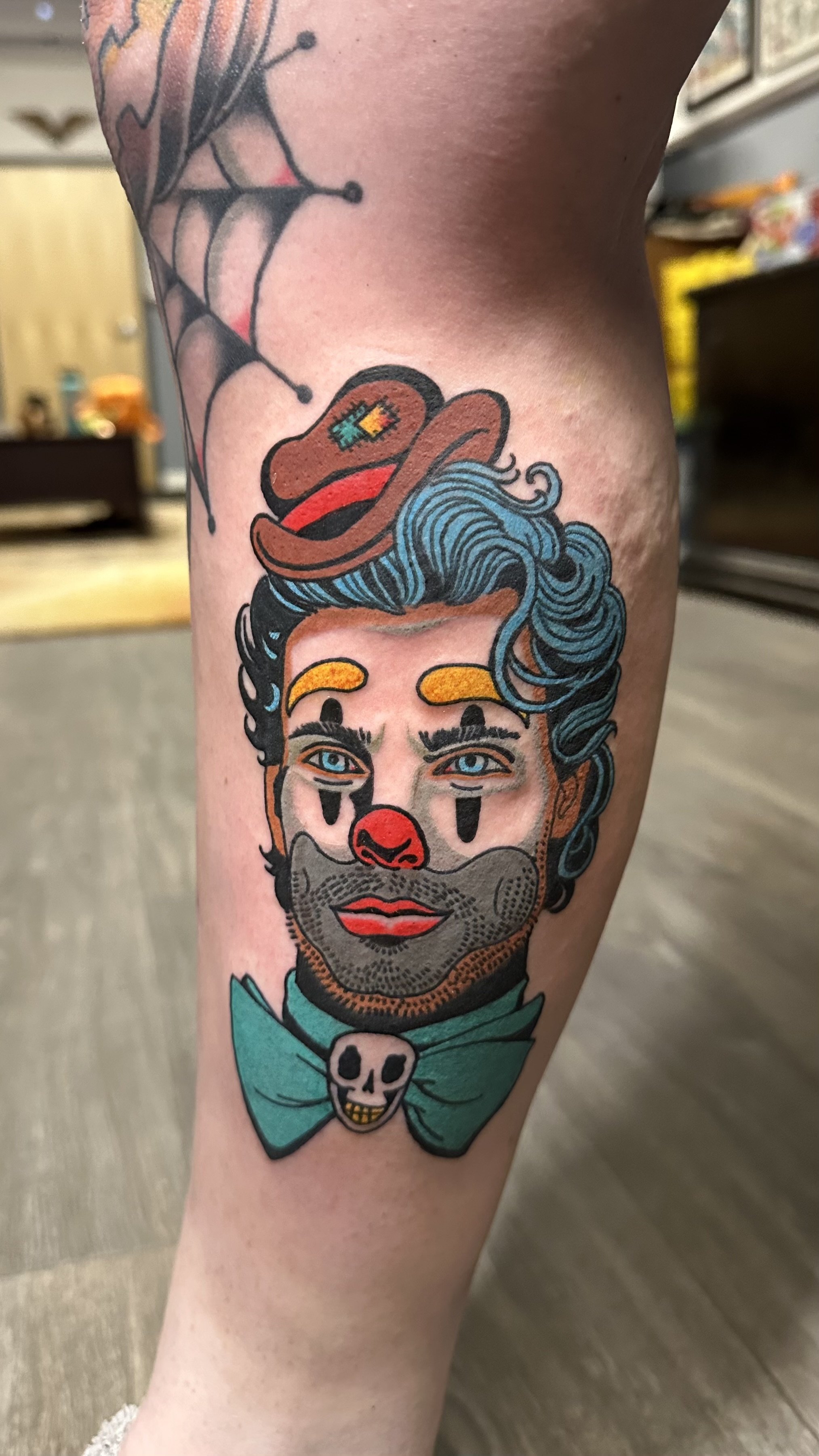 Tattoo of a man's face with blue hair, a mustache, and a skull on his shirt, wearing a clown nose and a hat with a pixel heart on it. A spider web is above his head.