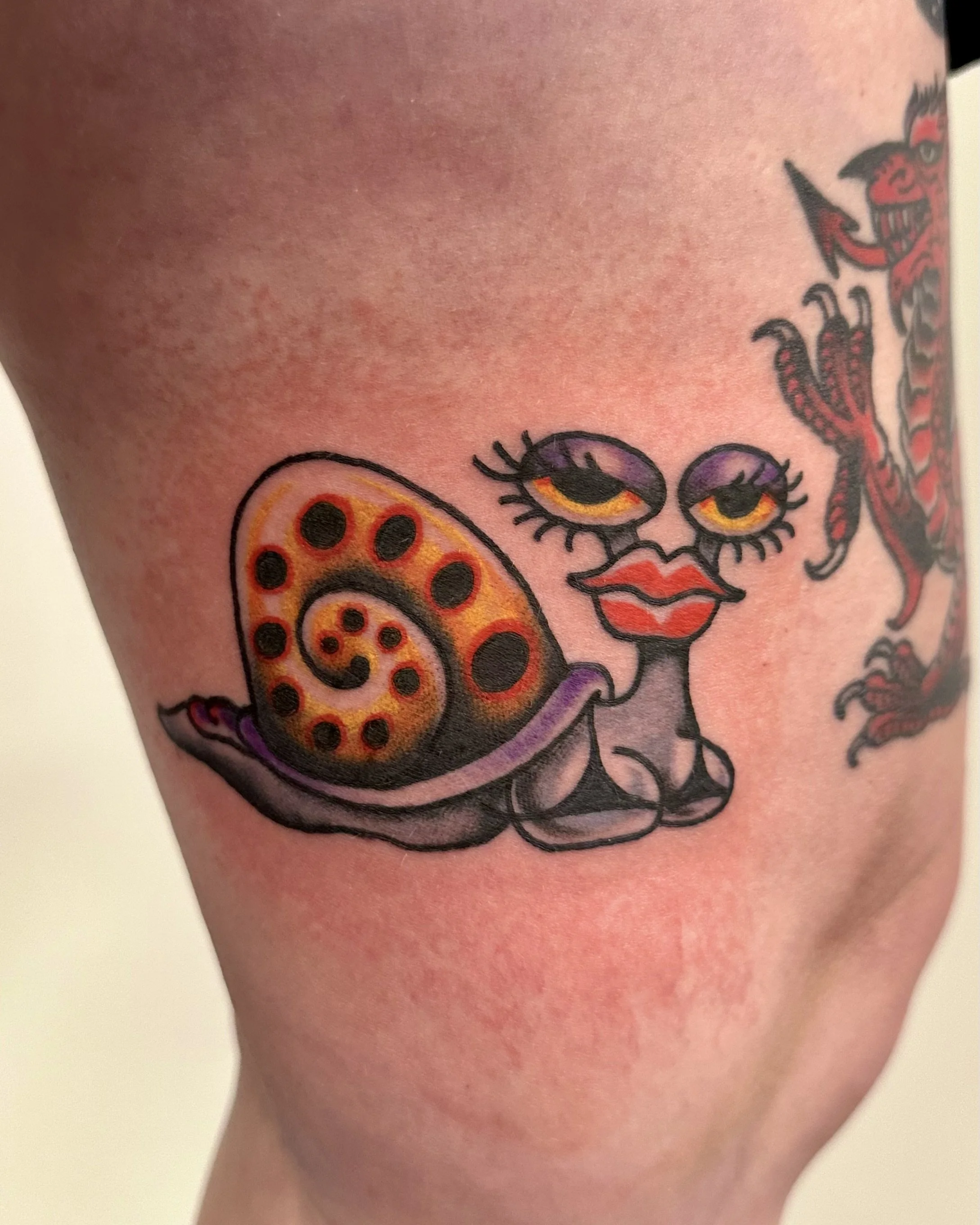 Close-up of a colorful tattoo on skin featuring a cartoonish ladybug with orange and black spots, expressive eyes, red lipstick, and a smiling face, along with a red devil with a mischievous grin and an arrow near its mouth.