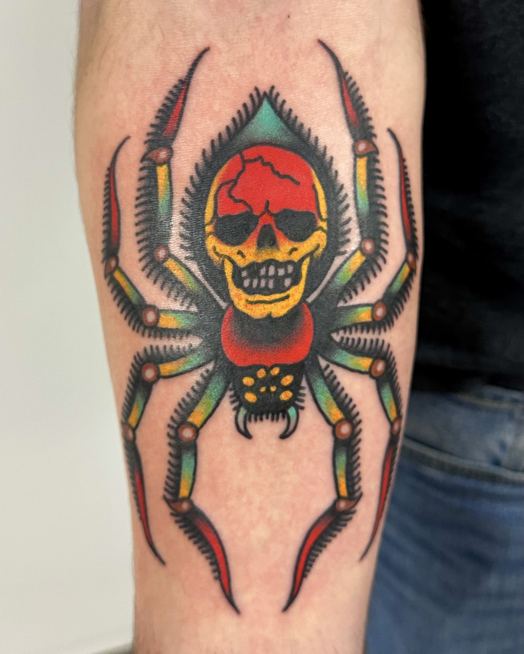 Tattoo of a red and yellow skull with black eye sockets and nose, surrounded by a spider with green, yellow, and red legs, on a person's upper arm.