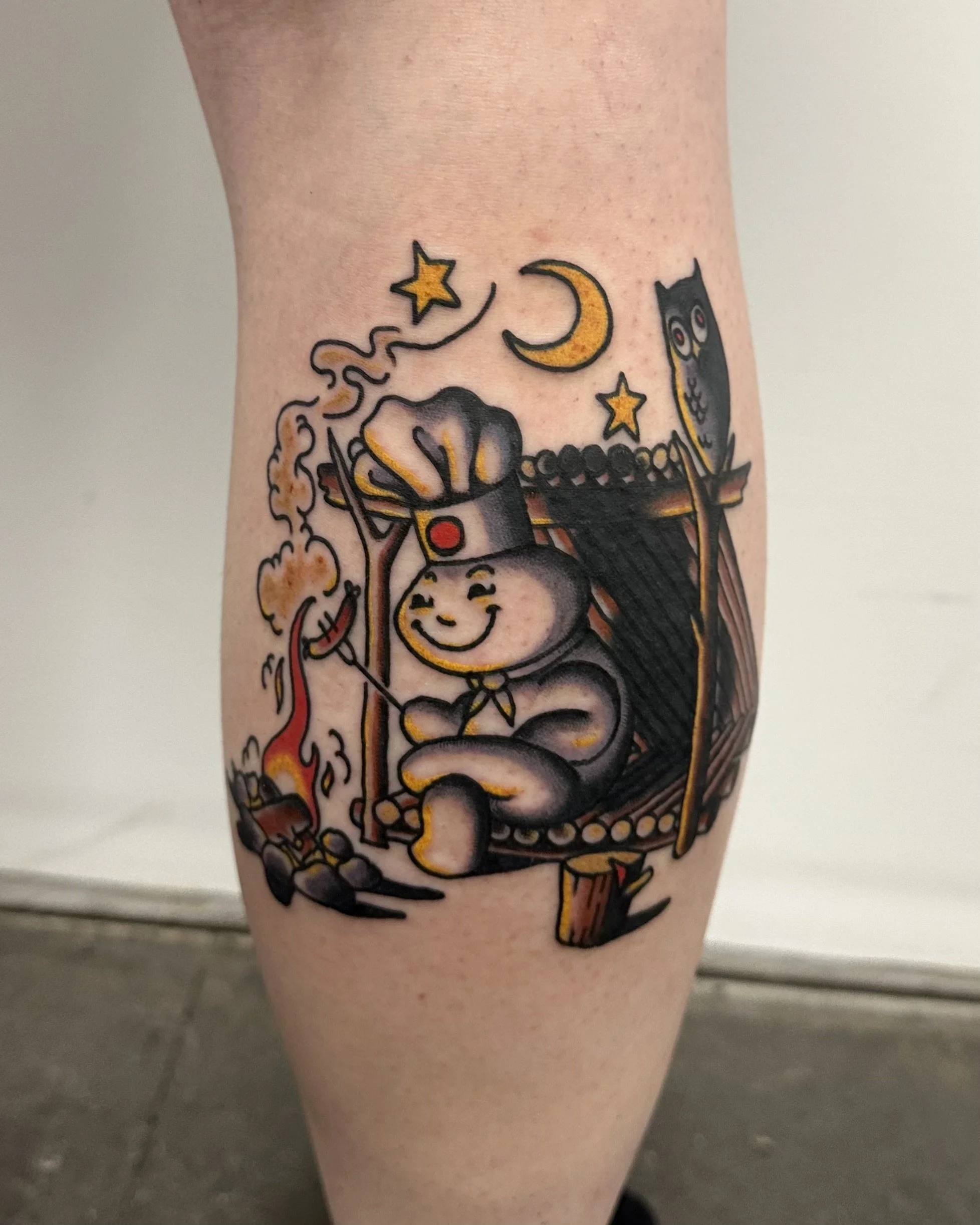 Colorful tattoo on a person's leg featuring a cartoon-style skeleton, flames, stars, a crescent moon, and an owl.