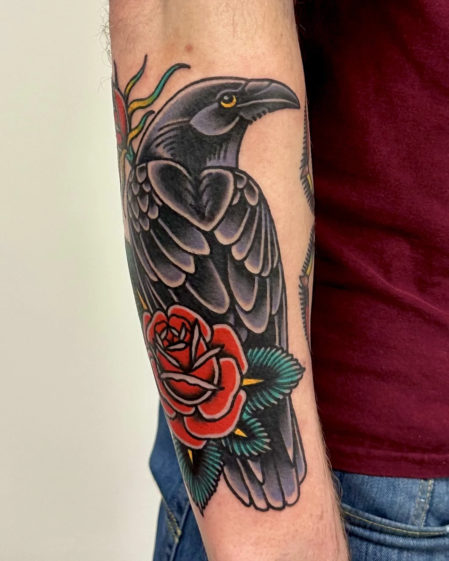 A colorful tattoo of a bird with black and gray feathers, yellow eyes, and colorful accents, intertwined with red roses and green leaves on a person's arm.