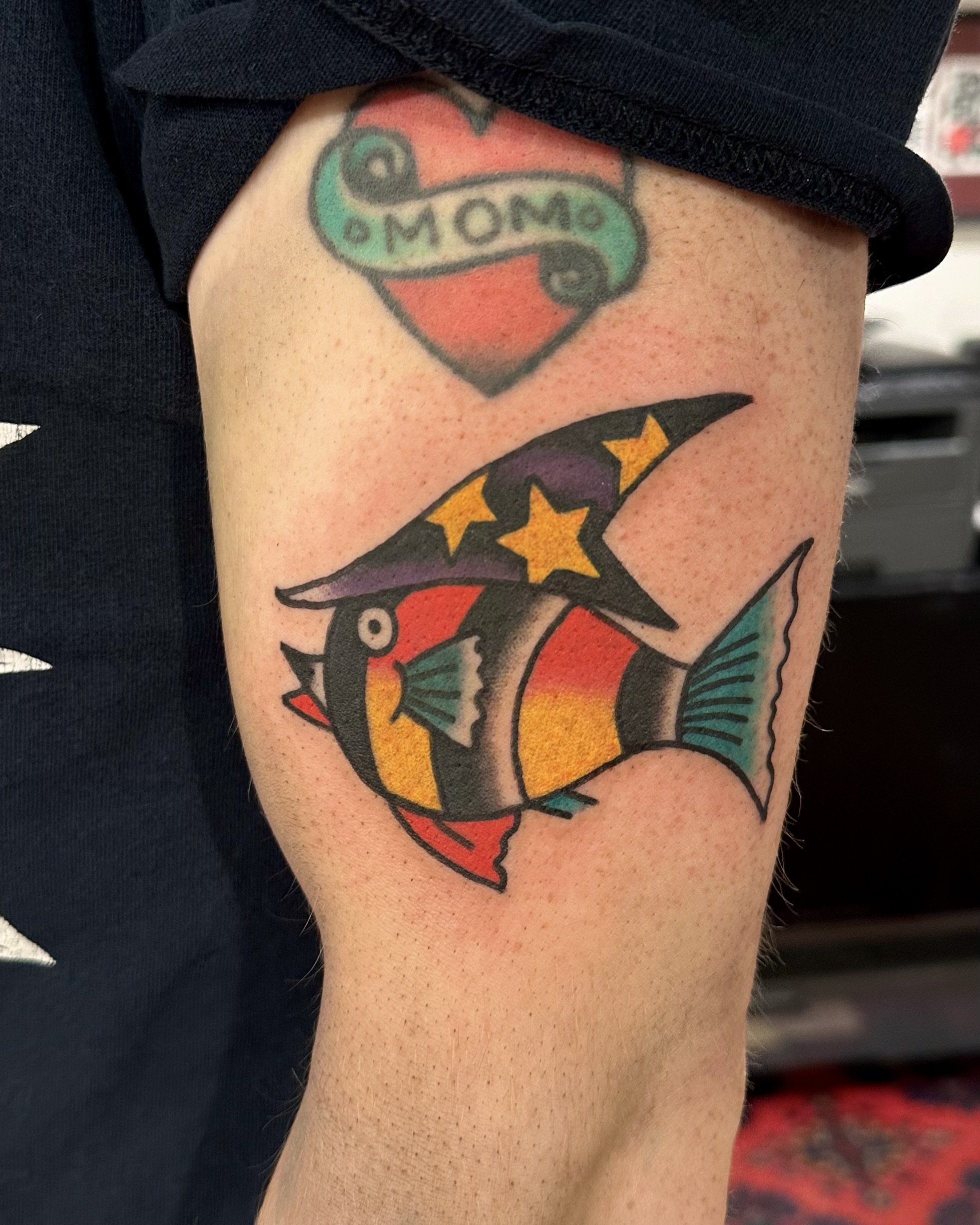 Colorful tattoo on person's upper arm featuring a bat with yellow eyes and orange and purple wings, with a red background and the word 'Mom' written inside a green banner.