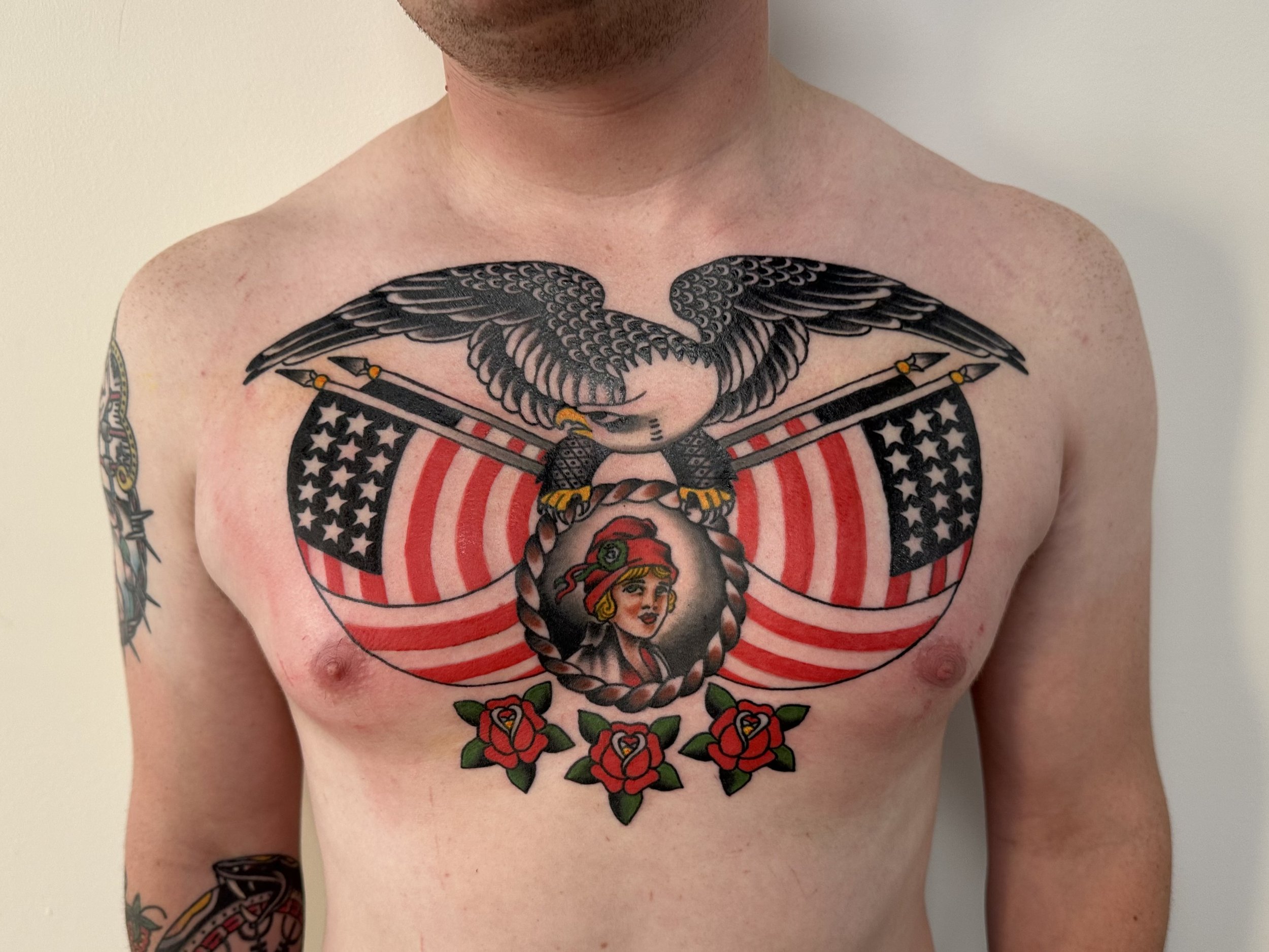 Partial view of a person's upper body with tattoos on chest and arm, featuring a large eagle, stripes, stars, a woman, and roses.