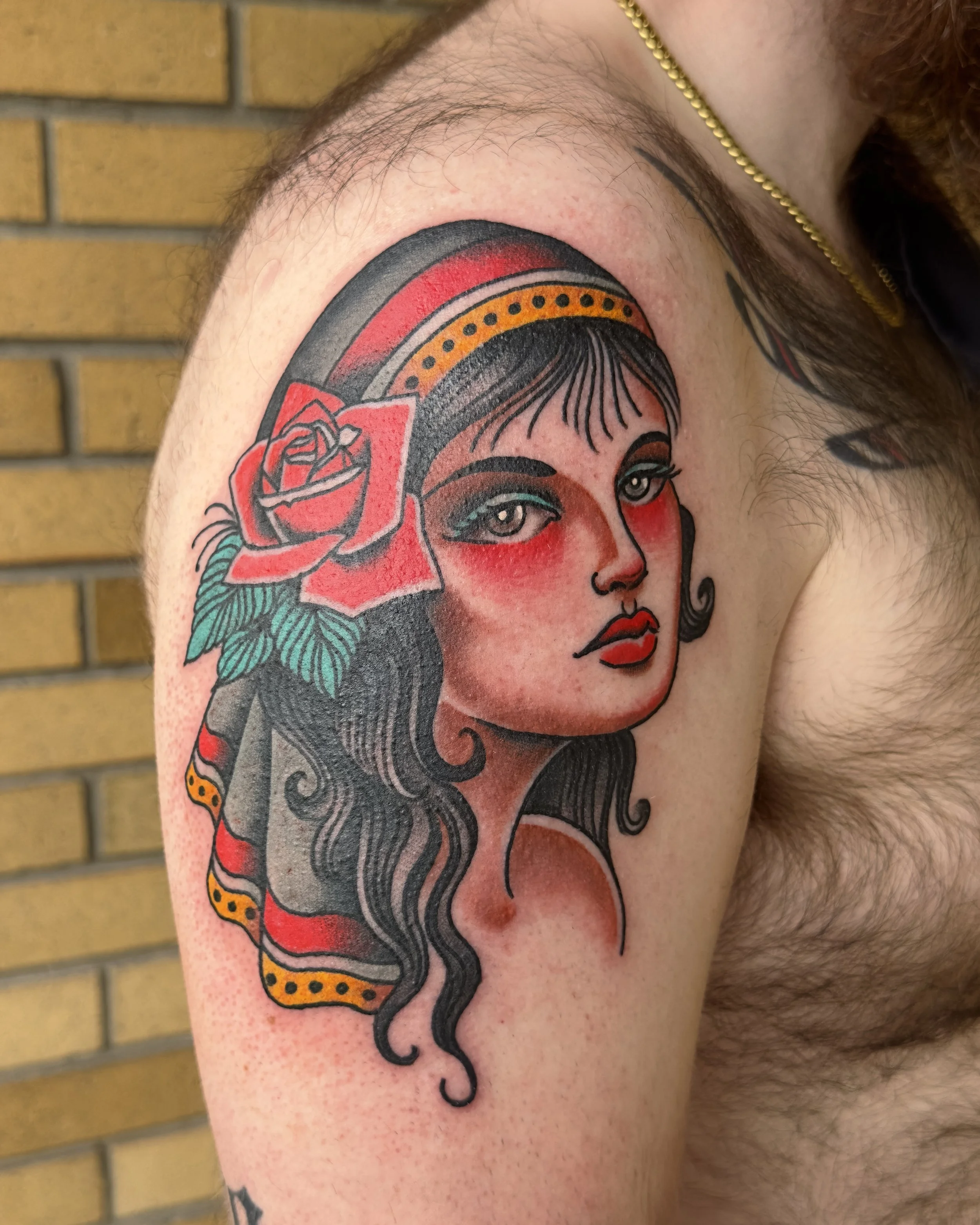 A tattoo of a woman with a rose in her hair on a person's upper arm, with a rainbow in the background, against a brick wall. The woman has light skin, blue eyes, and dark hair.
