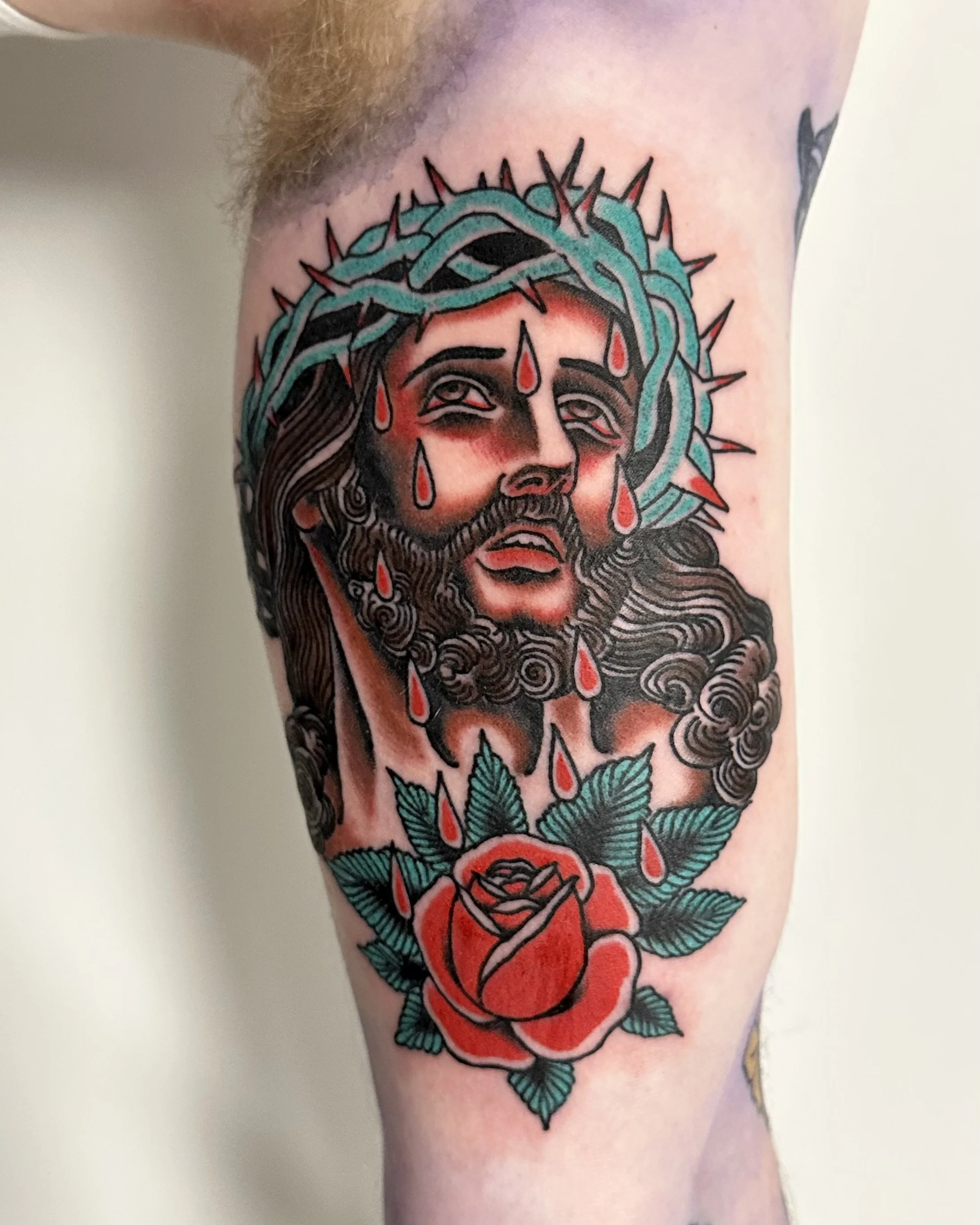 Tattoo of a woman’s face with a crown of thorns around her head, tears, and a rose at the neck on a person's arm.