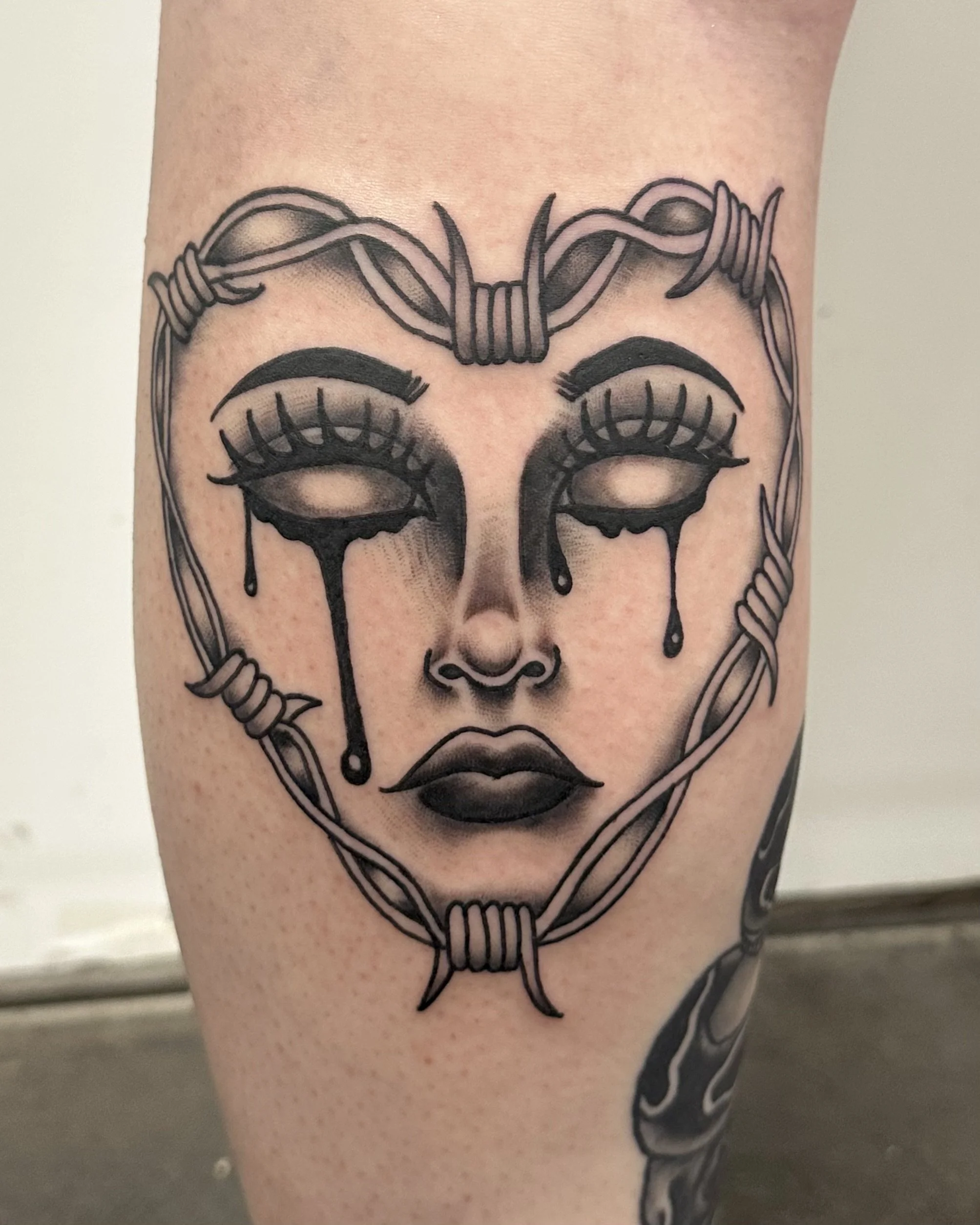 Tattoo of a stylized face with closed eyes, tears, and a nose ring, surrounded by a wire frame.