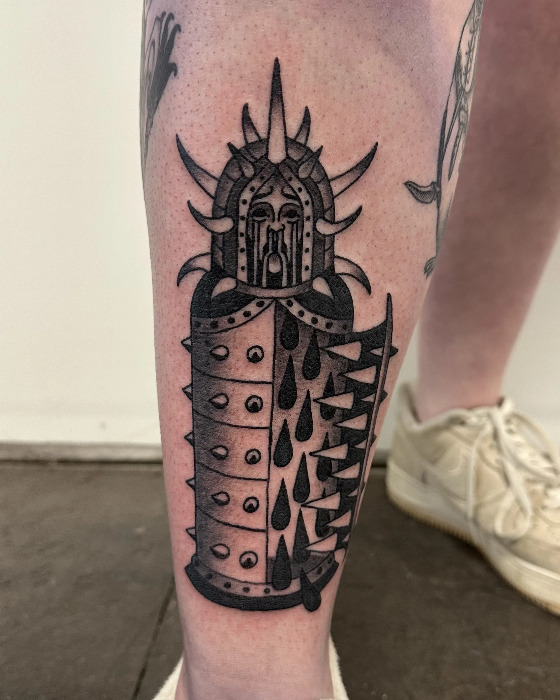 Black tattoo of a stylized dagger or sword with geometric and tribal design elements on a person's lower leg.