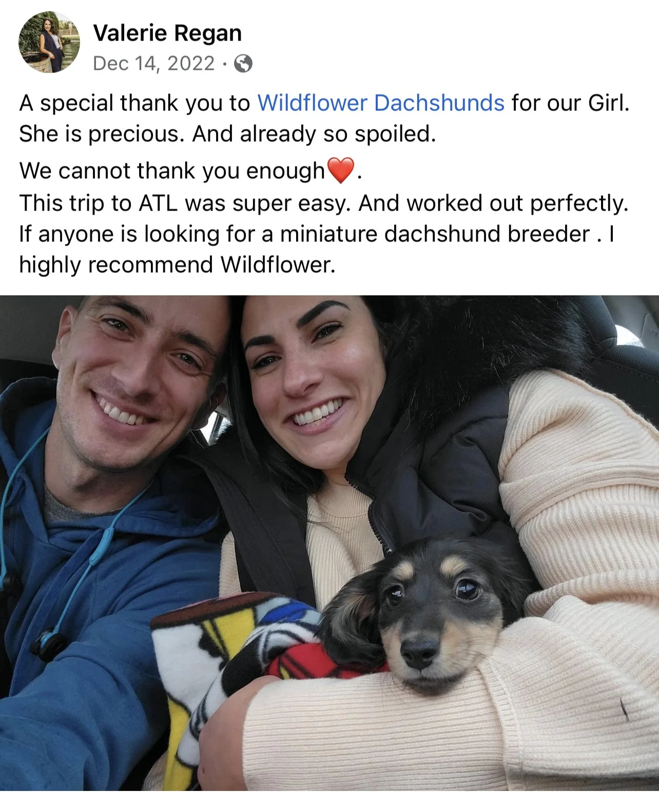 Two smiling people holding a small black and tan dachshund puppy wrapped in a blanket inside a vehicle.
