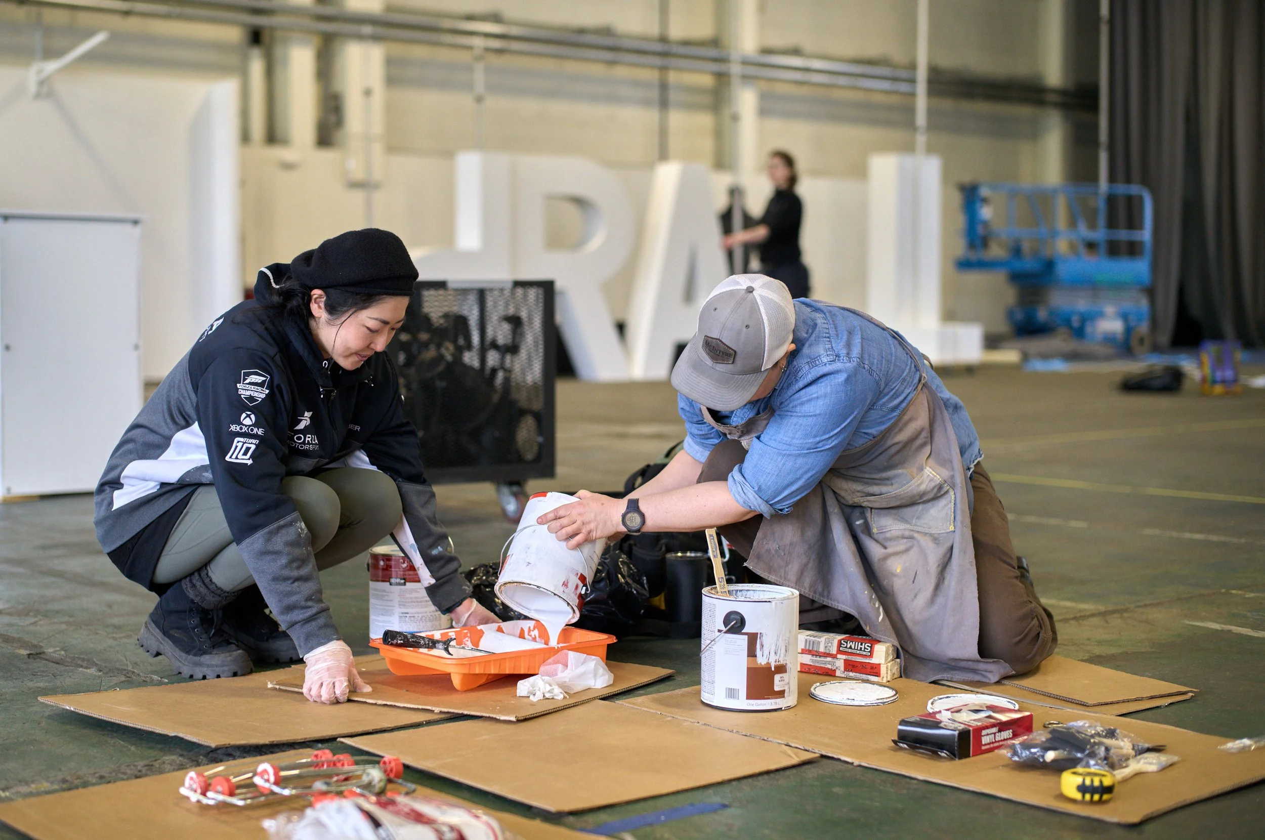 Two people are kneeling, pouring paint into a roller tray. They are surrounded by tools and supplies.