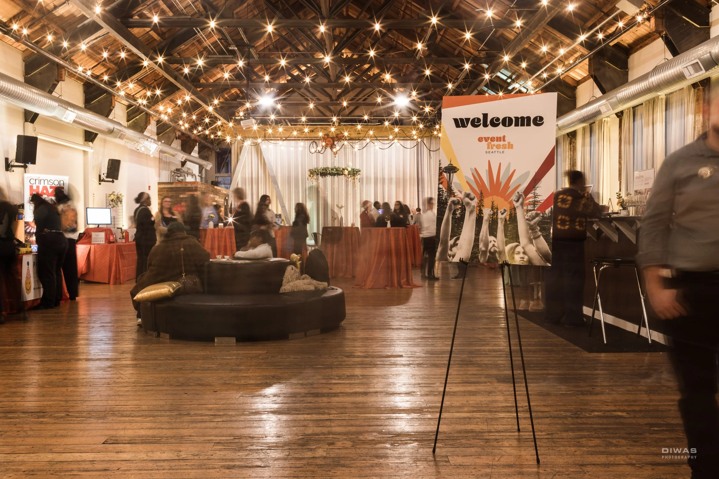 A motion blur of guests mingling at an event with cocktail tables, a bar, and a low seating circular sofa.