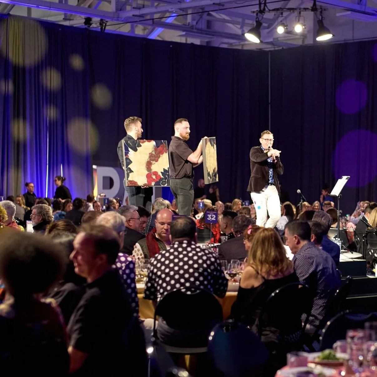 A room full of event guests seated at dining tables, and onstage there are two people holding artwork and an auctioneer pointing into the crowd.