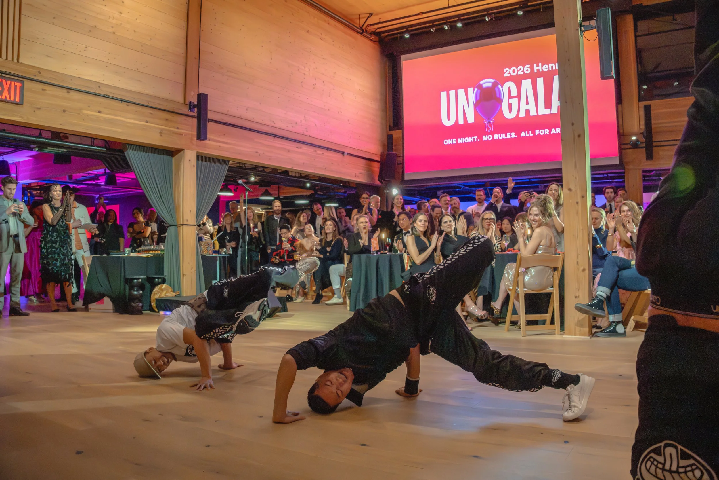 Two breakdancers are tilted dancing while guests are watching and cheering behind them.