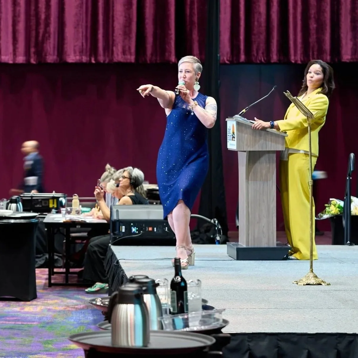 A woman in a blue dress points towards the crowd while another woman in a yellow suit is by the podium looking over. 
