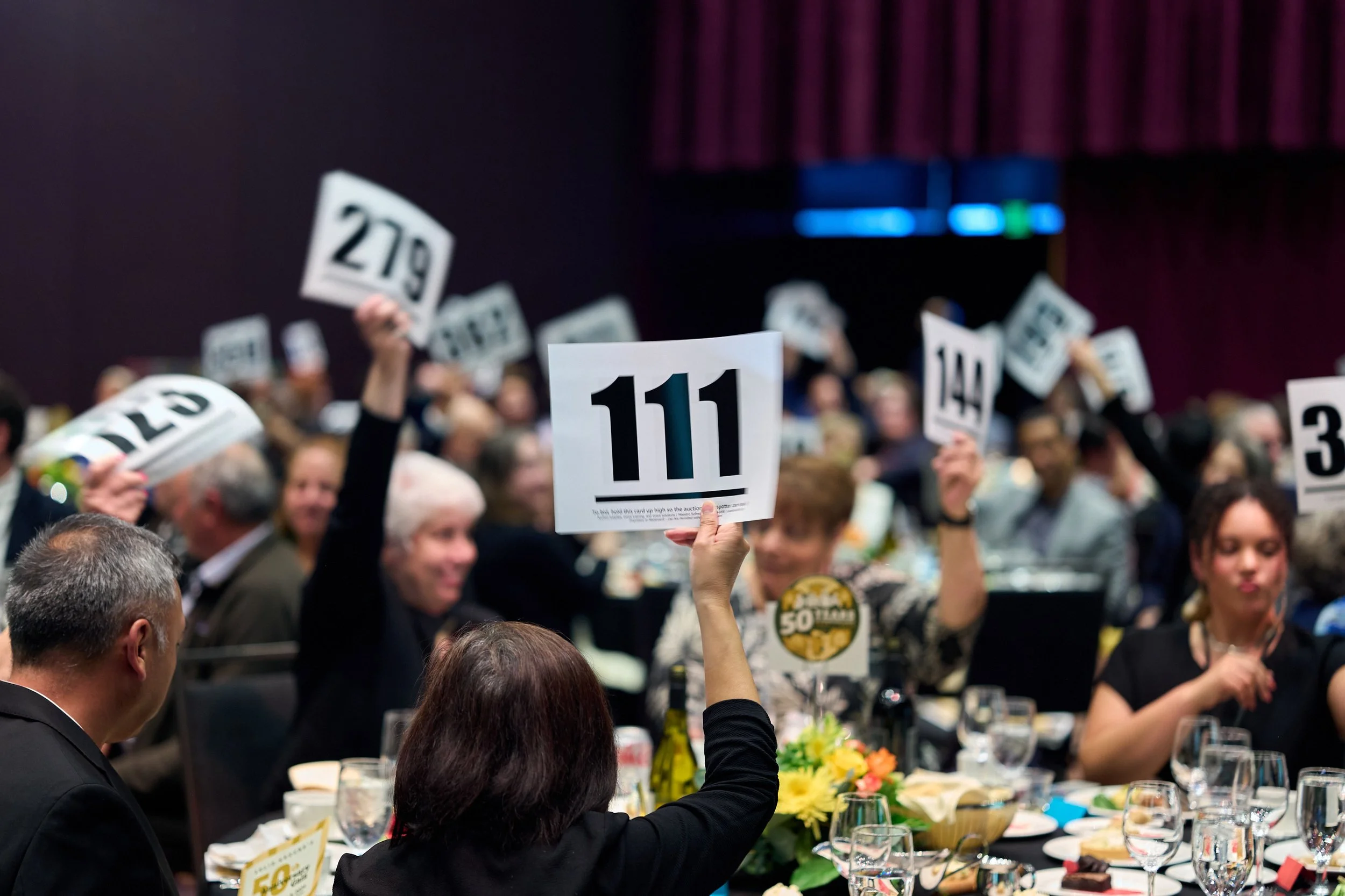 Guests are seen raising their bid numbers while sitting on round dining tables.