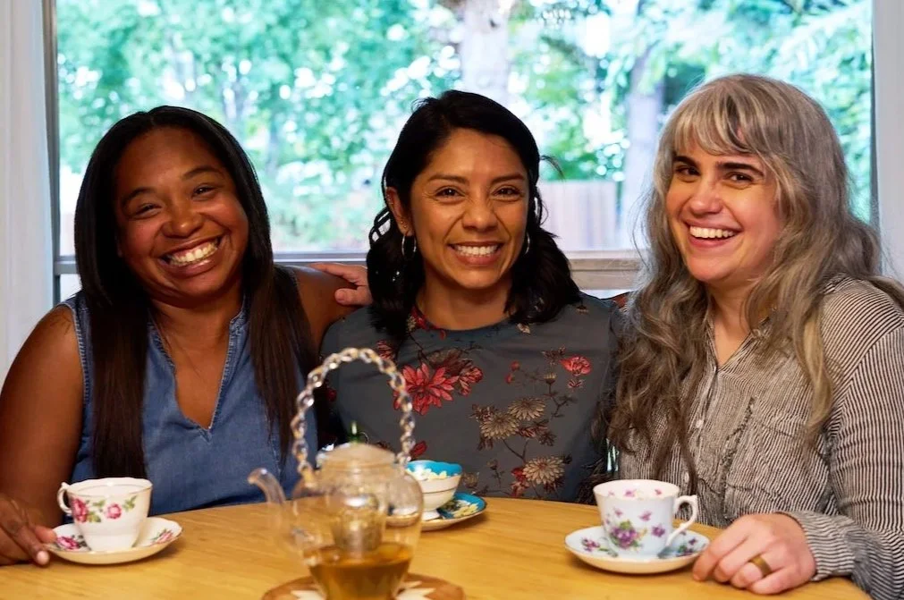 Three women smile while seated at a table, with colorful teacups and a teapot.