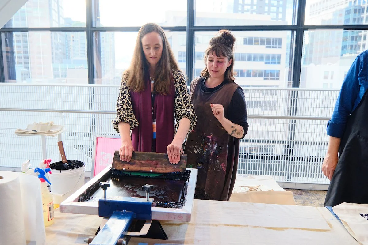One woman is creating a silk screen printing while the other woman with an apron looks over to guide her with the process.