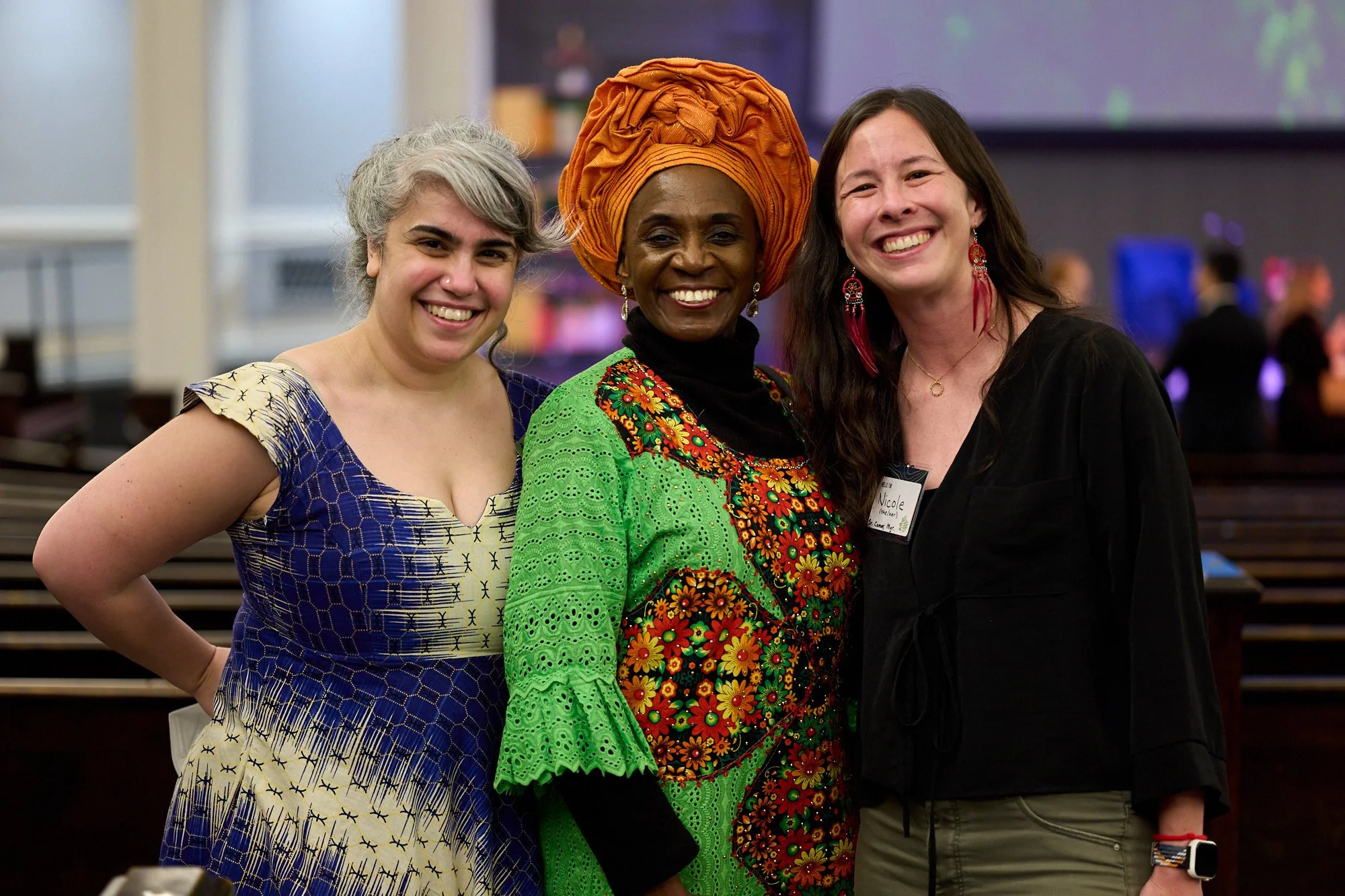 Three women are smiling at the camera while some guests are seen behind mingling.