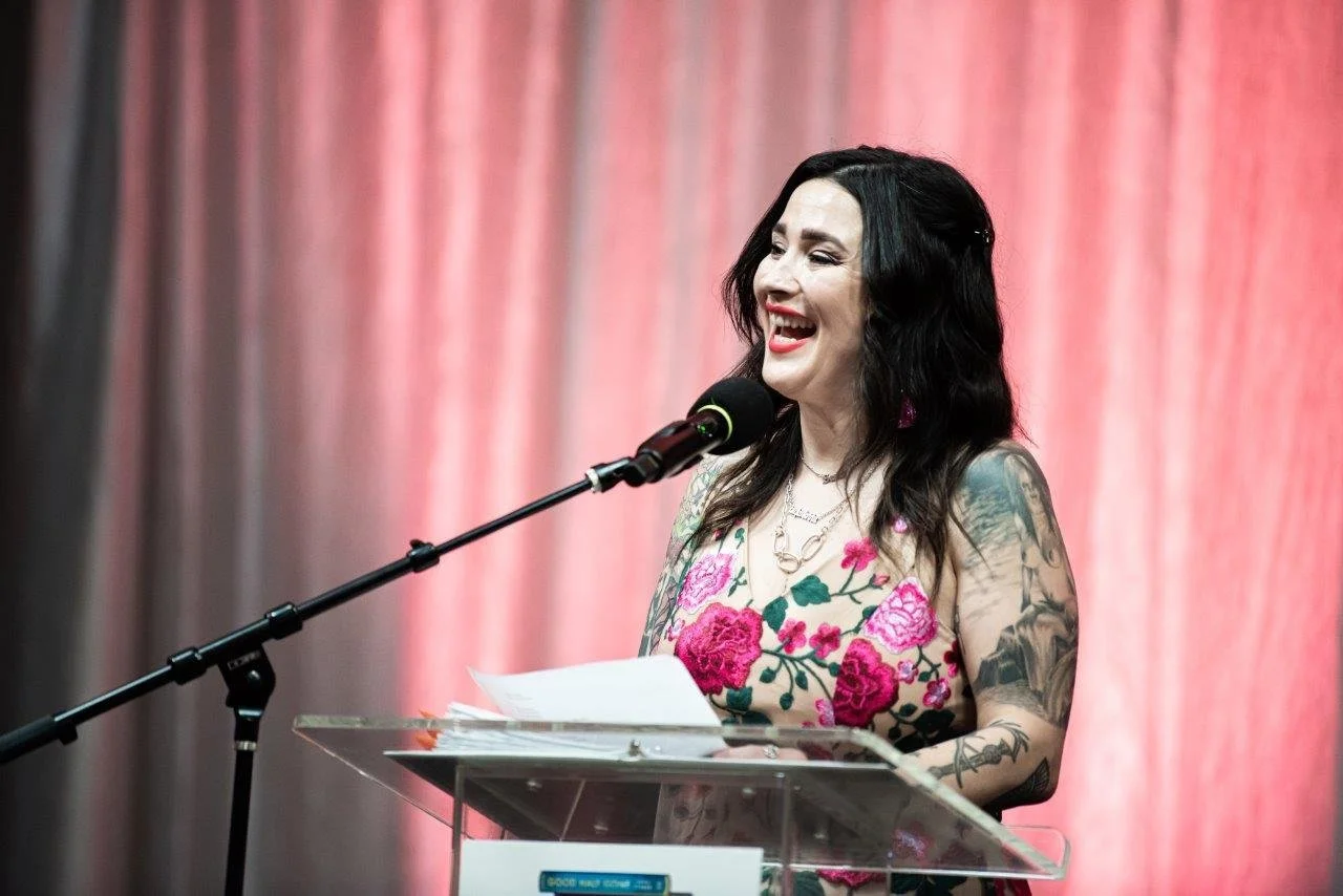 A dark-haired white woman wearing a pink floral dress  is joyfully speaking on a podium