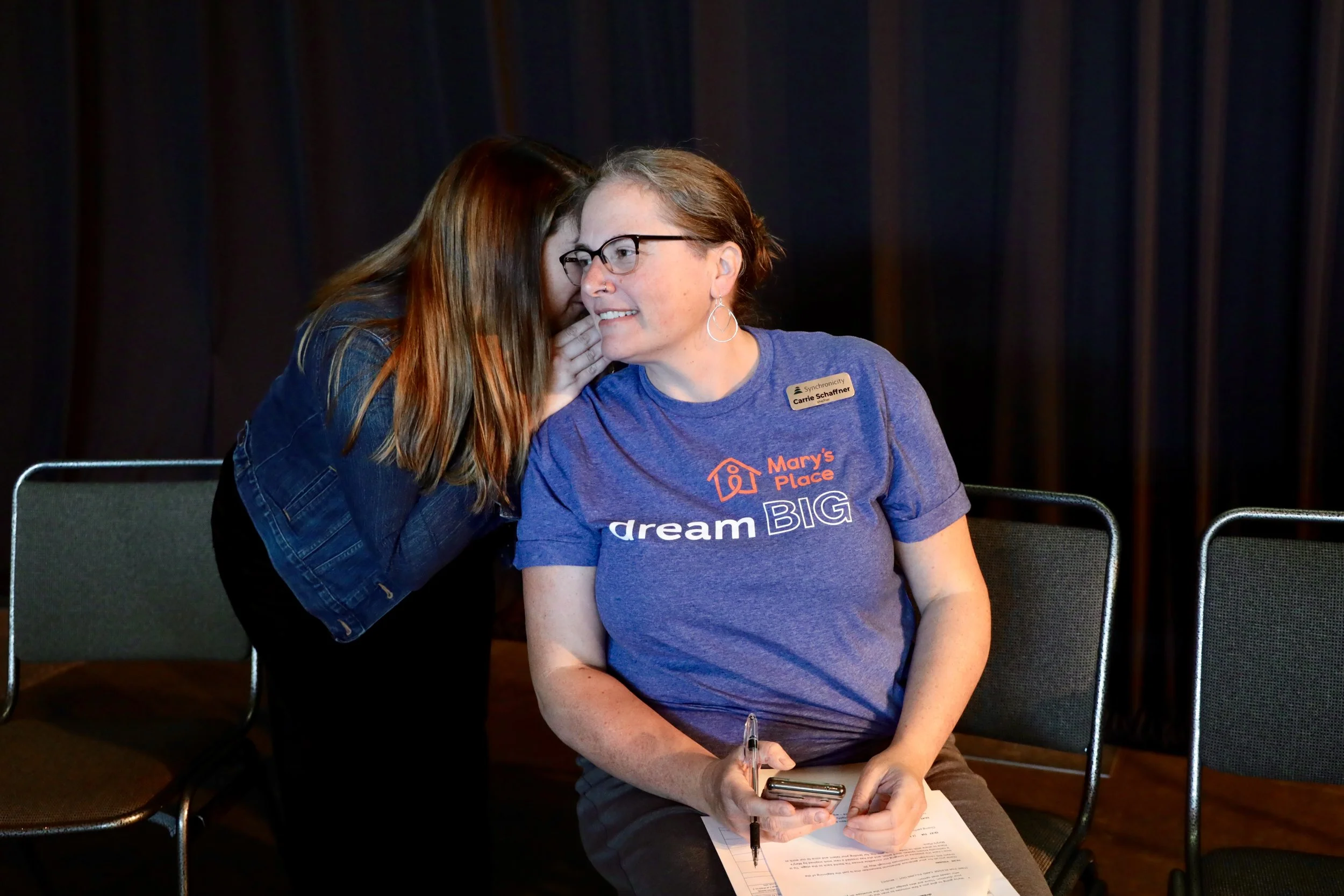 A woman is seated with her phone, pen and documents while another woman is whispering in her ear.
