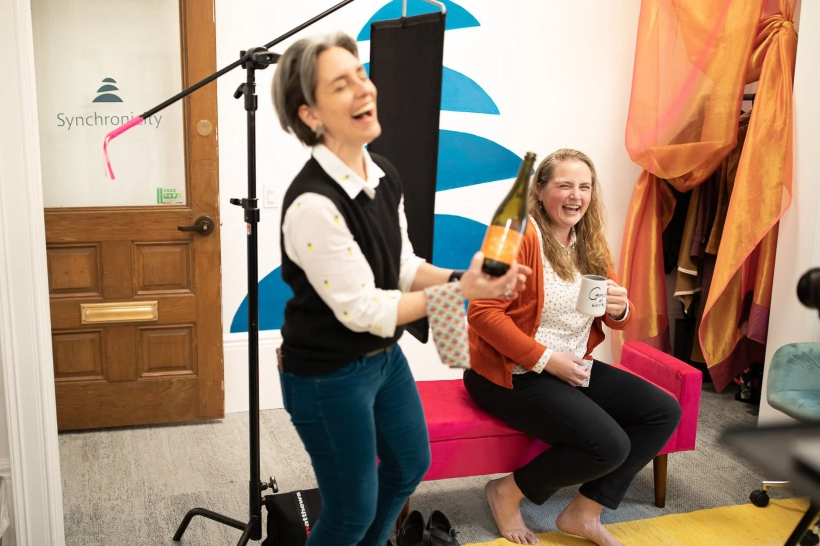 Two women are laughing in a colorful office setting. One holds a champagne bottle, the other holds a mug.