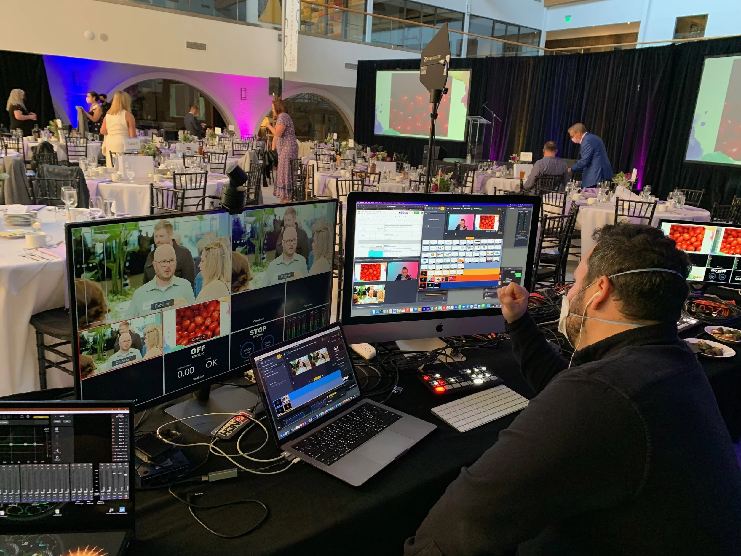 A person sits at a tech station facing two large monitors. In the background we see a room with dining tables and a few guests arriving.