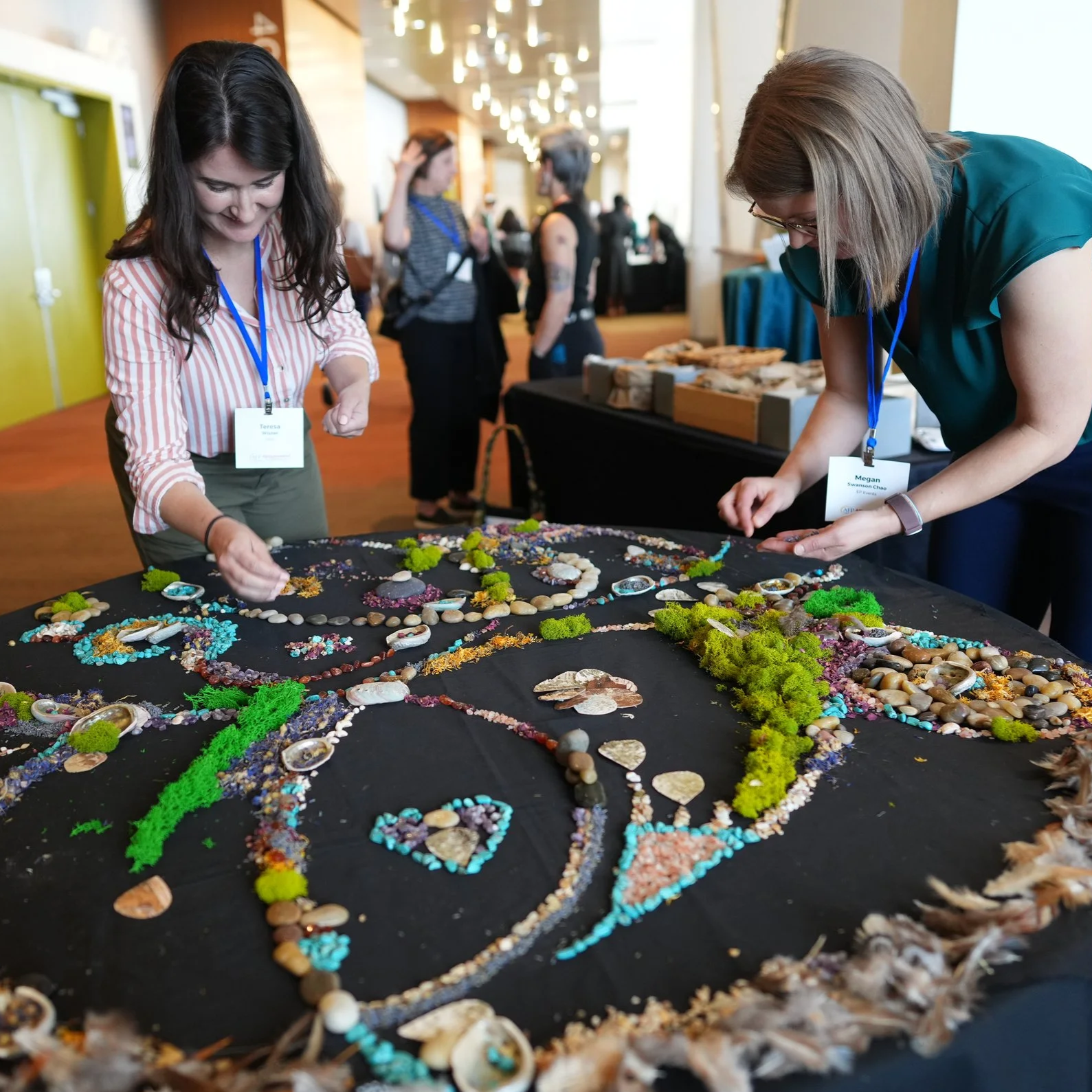 Two women are leaning over a table that has different textured pieces such as rocks, grass, pebbles, shells to create a collaborative art piece table. 